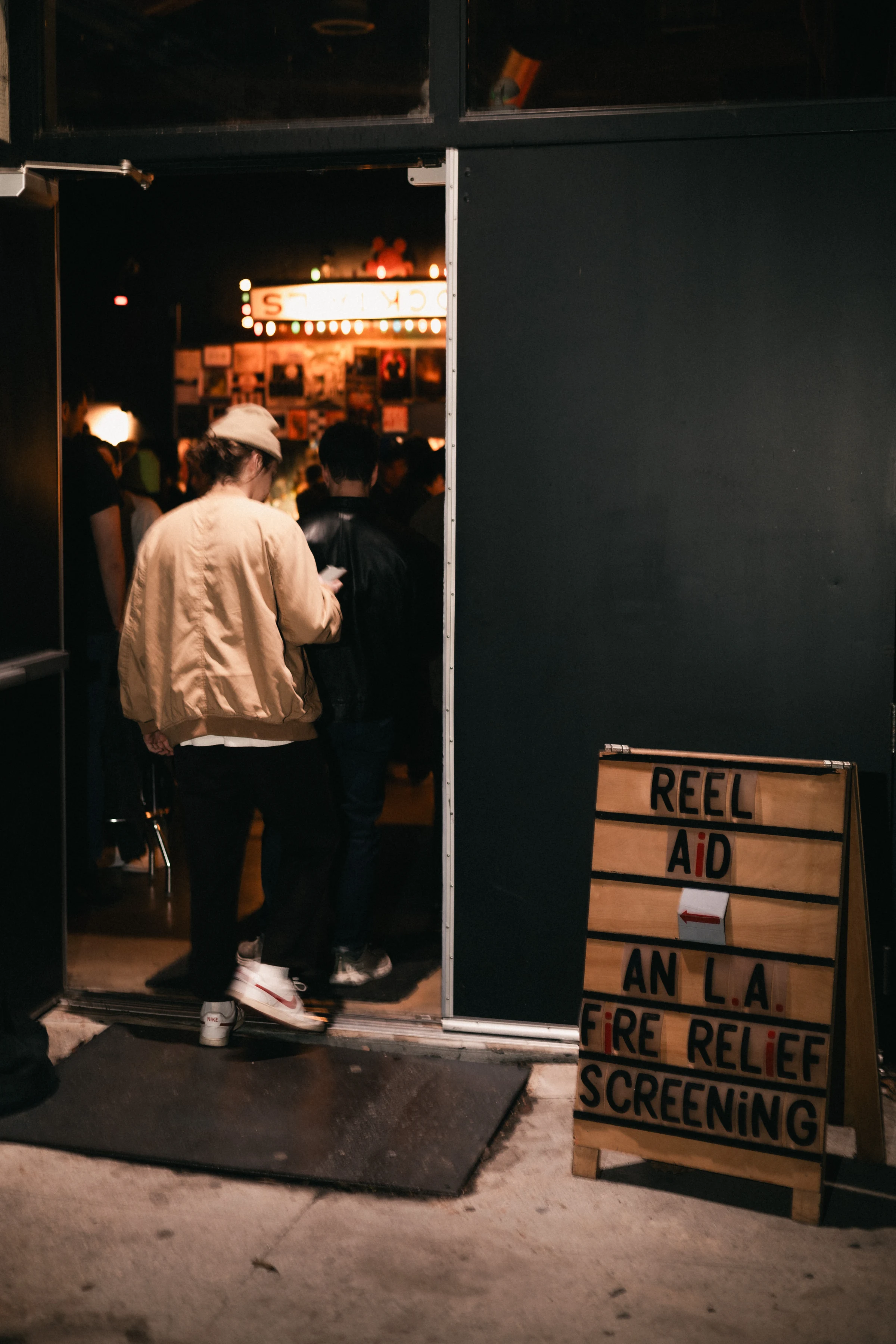 Person in a tan jacket and cap enters a dimly lit venue. A wooden sign reads "Reel Aid: An L.A. Fire Relief Screening" beside the door.