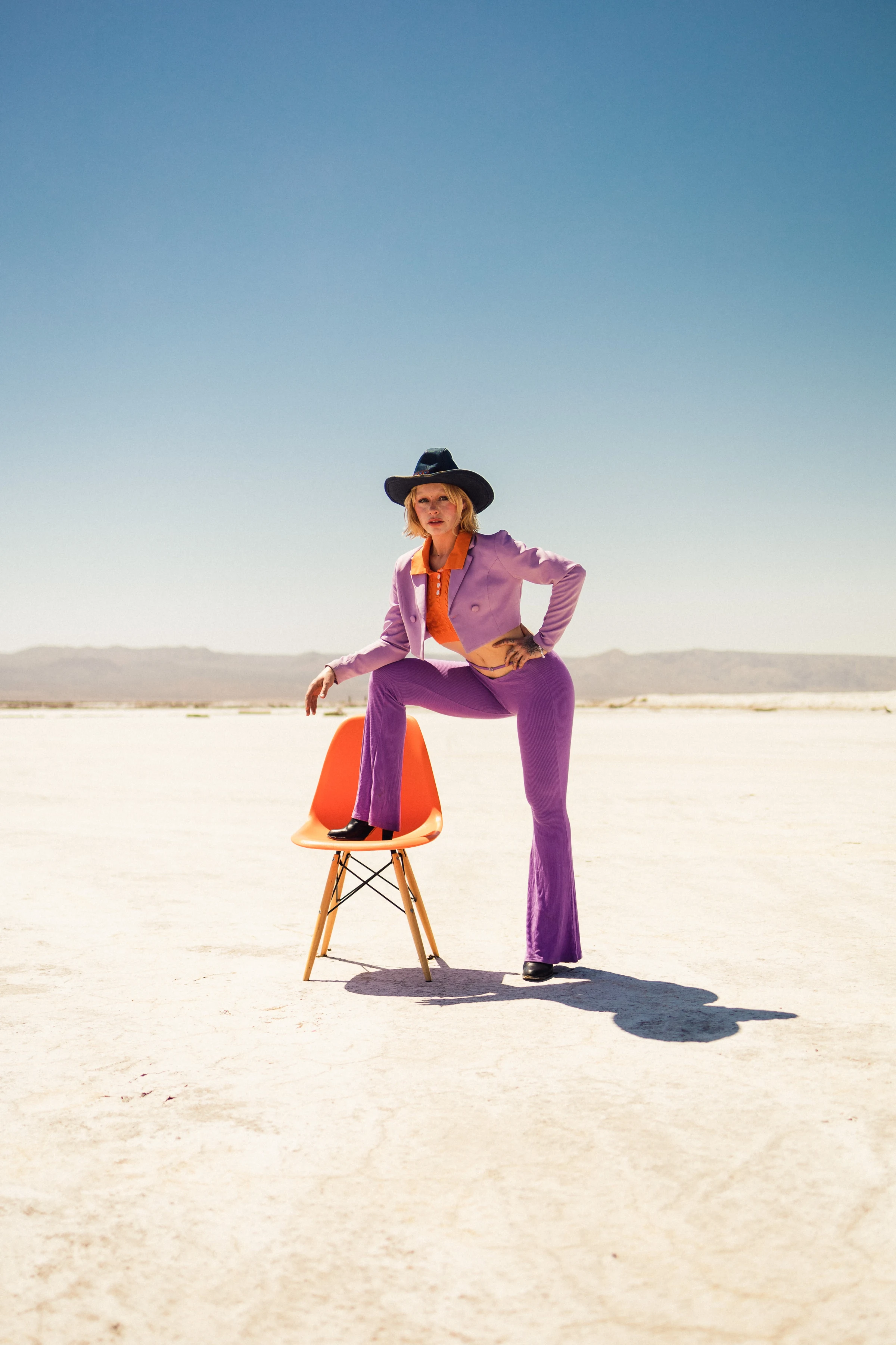 A woman in purple suit and black hat poses confidently with one foot on an orange chair in a desert, under a clear blue sky.