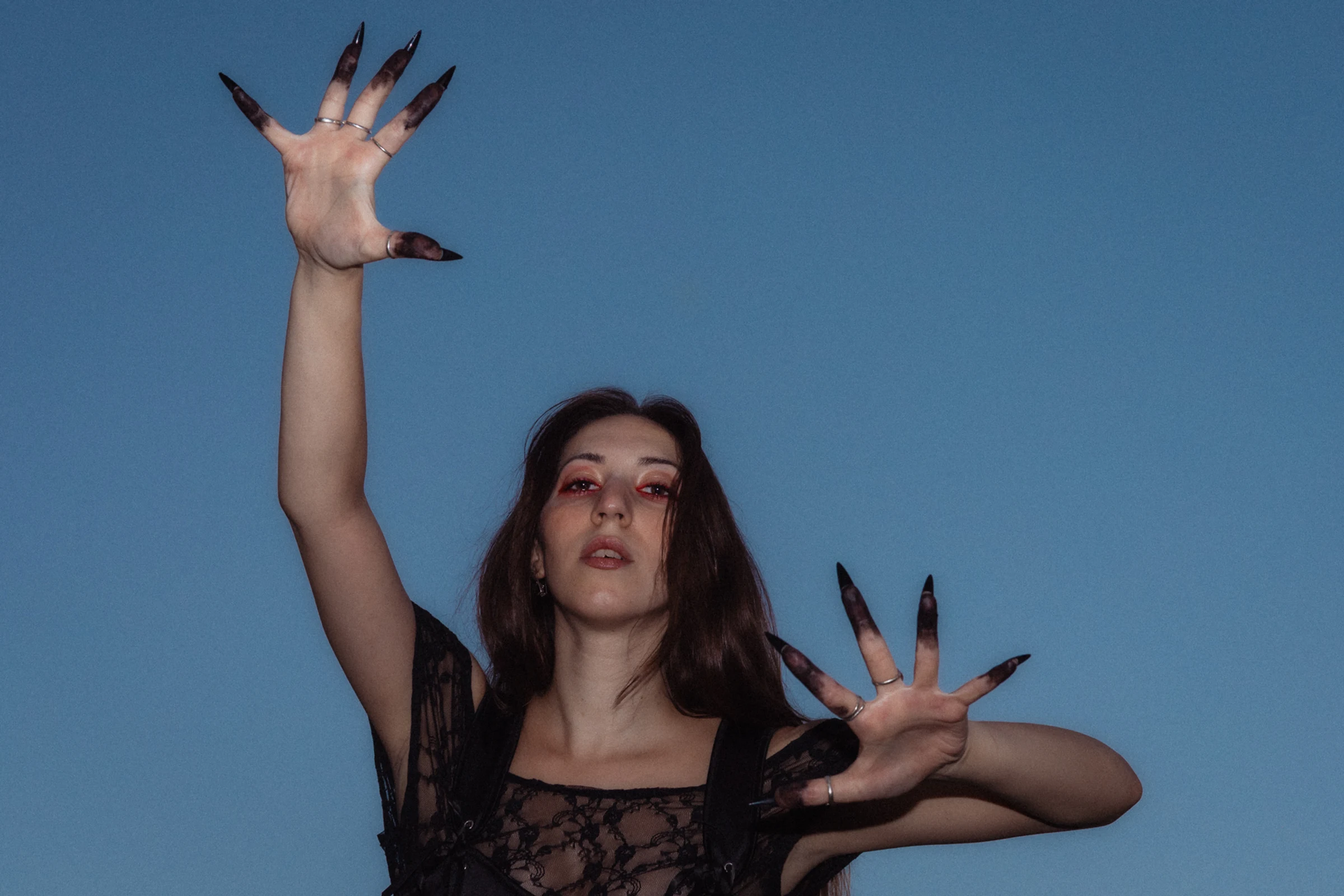 A woman with long dark hair and dramatic black claw-like nails poses against a blue sky. She wears a black lace top, her expression intense. Tree branches frame the scene.