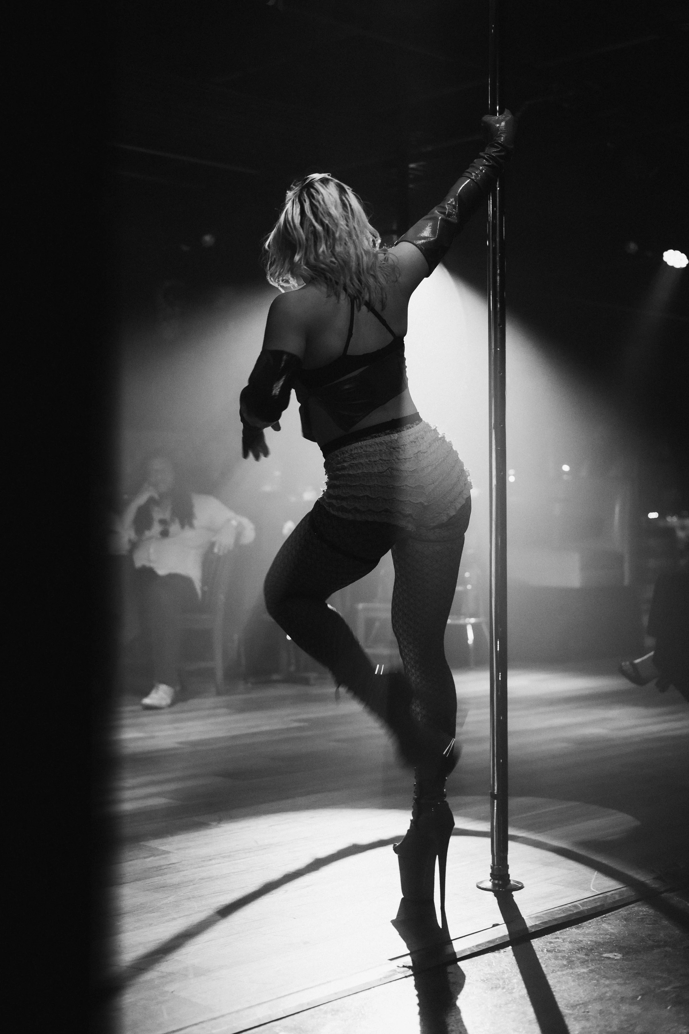 A woman is performing a pole dance on stage in dim lighting, wearing high heels and form-fitting attire. The atmosphere is moody and dramatic.