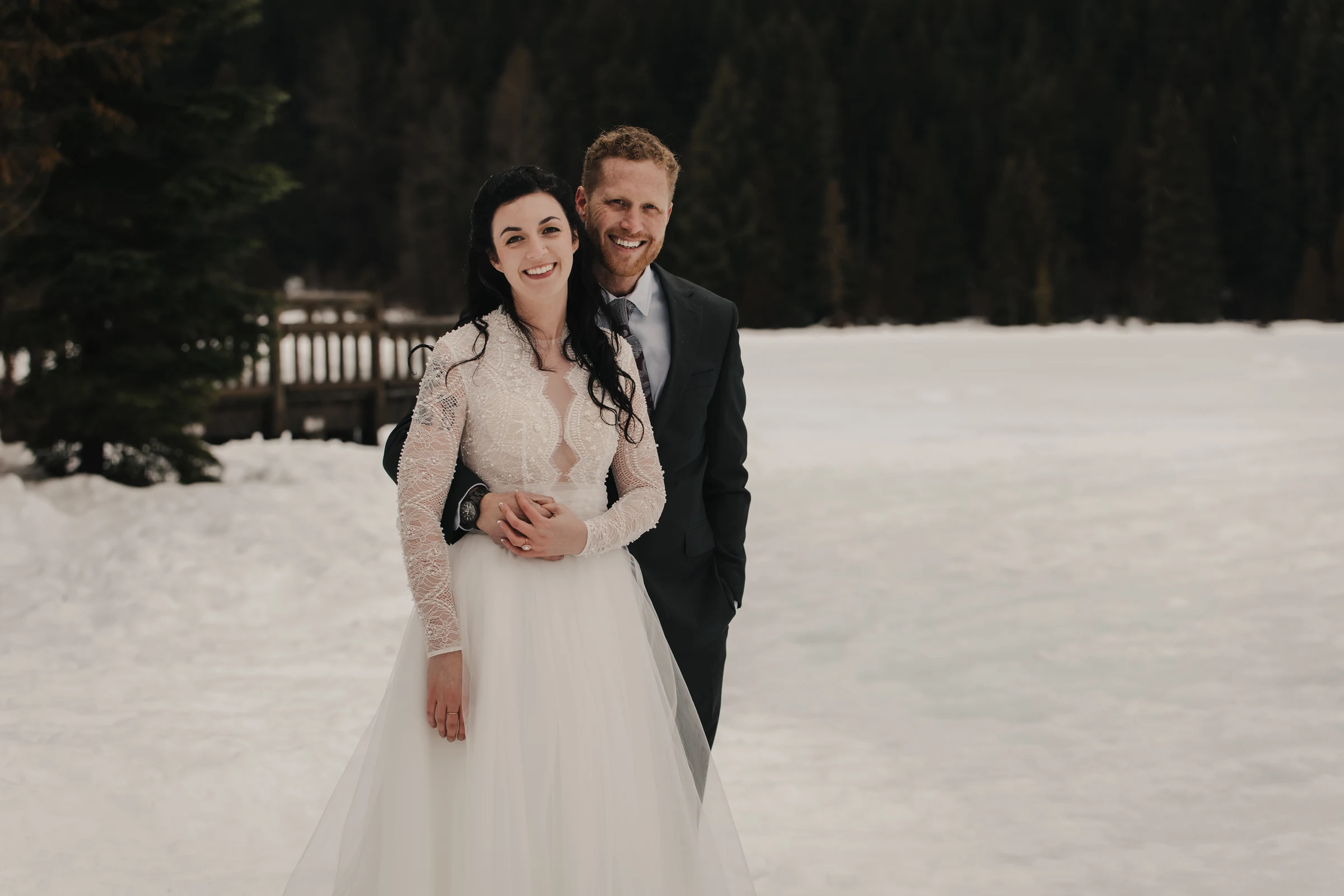 A smiling couple in wedding attire stands on snowy ground. The bride wears a lace gown, and the groom is in a suit. Forest trees form the background.