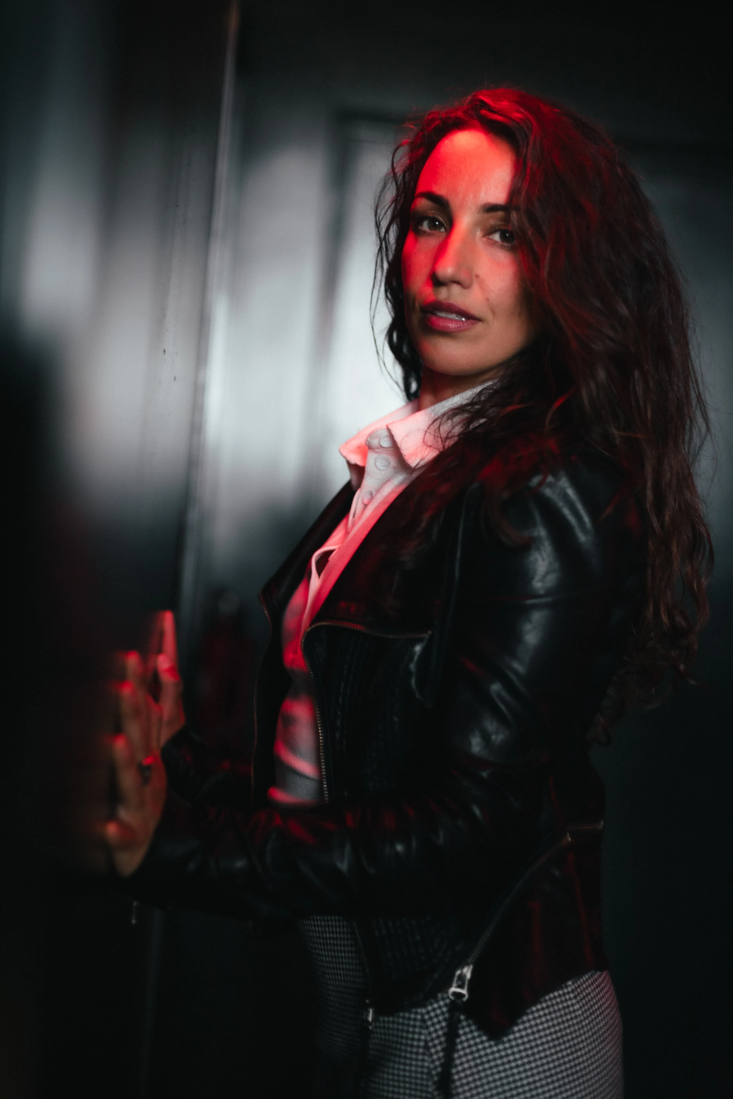 A woman with wavy hair, wearing a leather jacket and checkered pants, leans against a wall. Red lighting creates a dramatic and intense mood.
