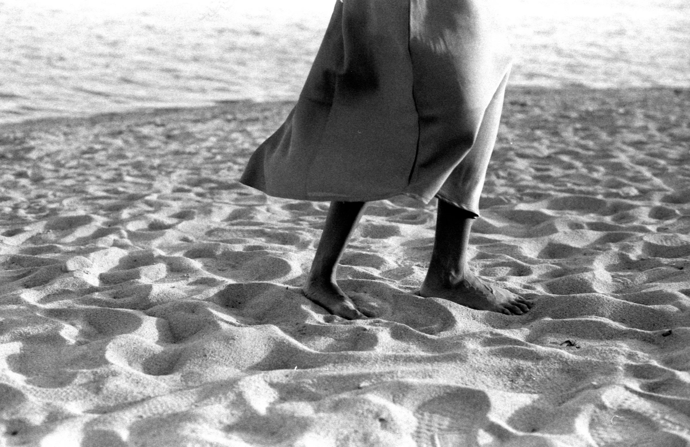 A person walks barefoot on rippled sand, wearing a flowing skirt. The black-and-white image captures a serene, contemplative beach atmosphere.