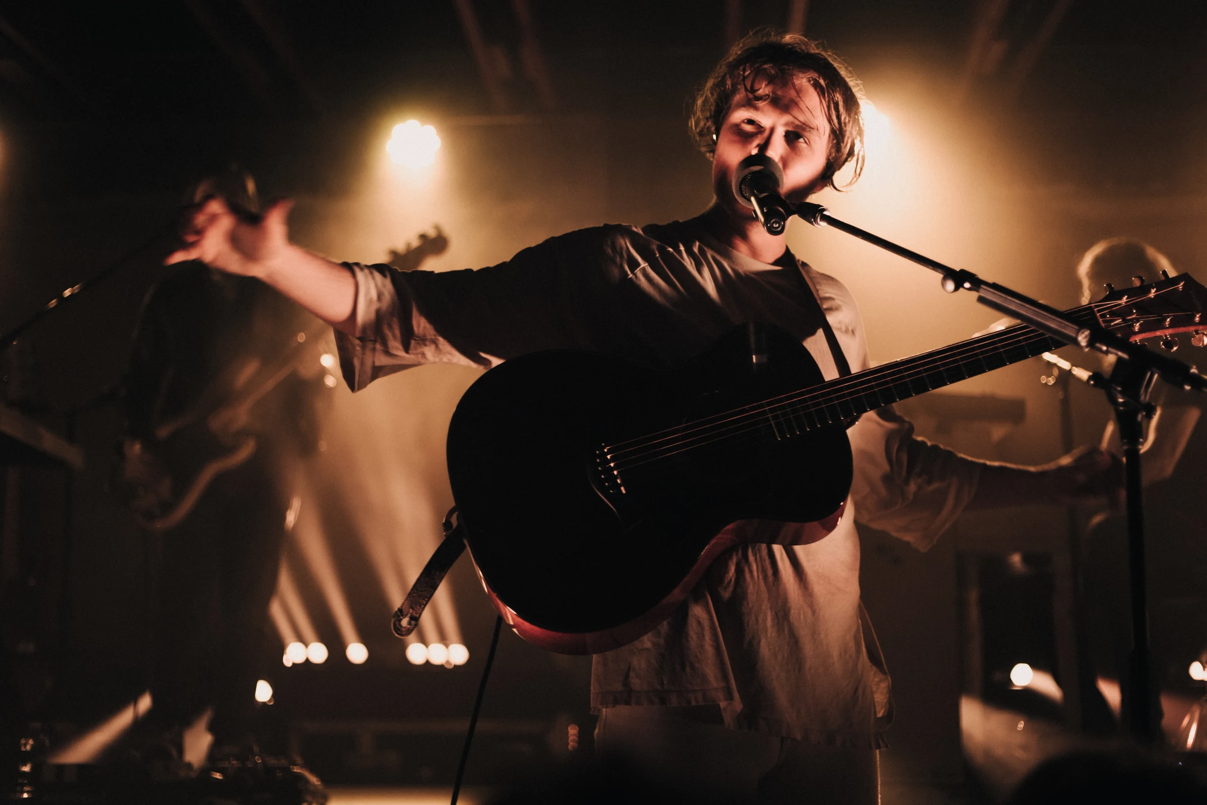 A musician passionately performs on stage with a guitar, illuminated by warm spotlights. The dimly lit background features band members playing instruments.