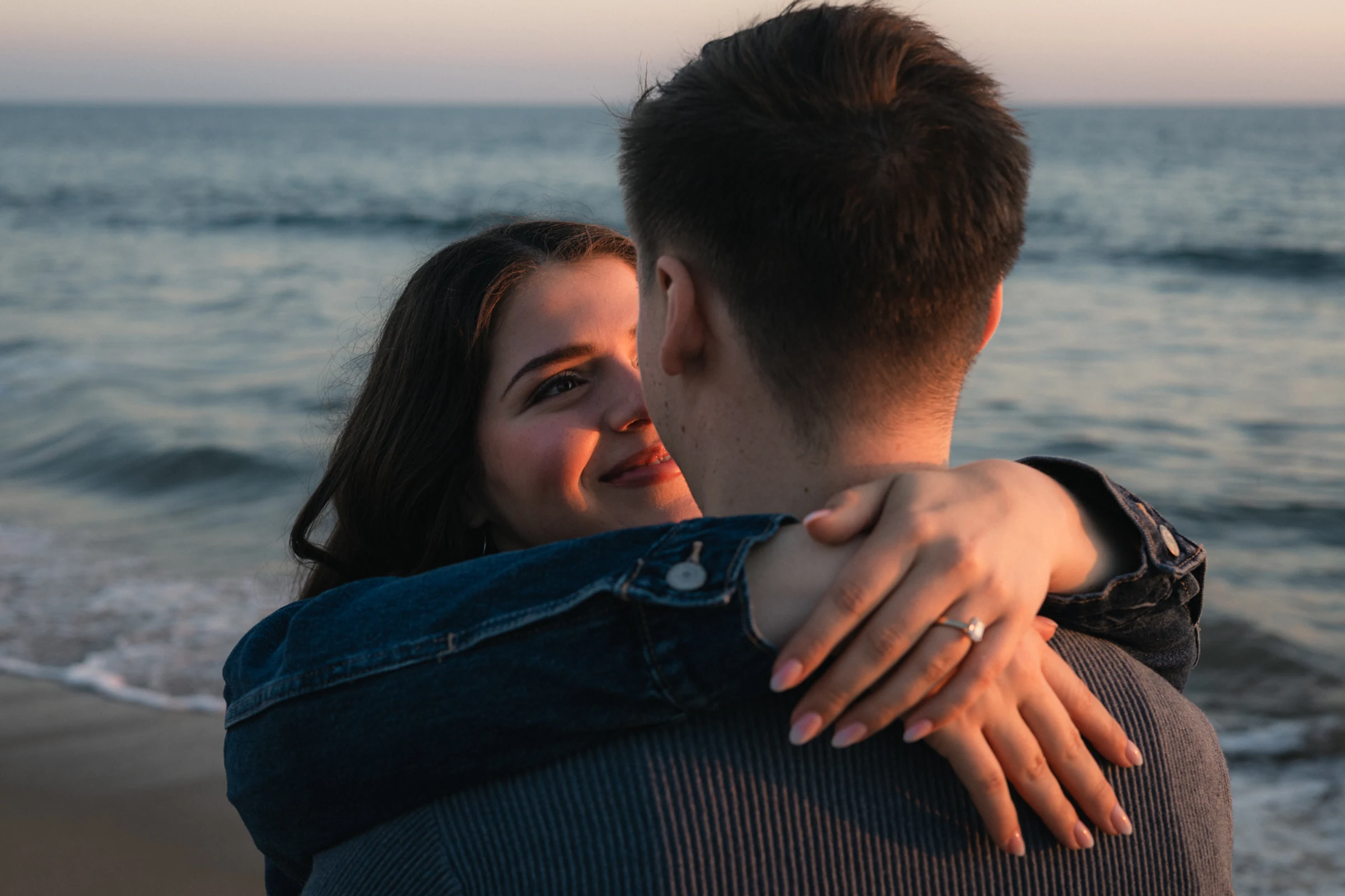 A couple embraces on a beach at sunset. The woman, smiling gently, wraps her arms around the man, who faces the ocean. A serene, romantic moment.