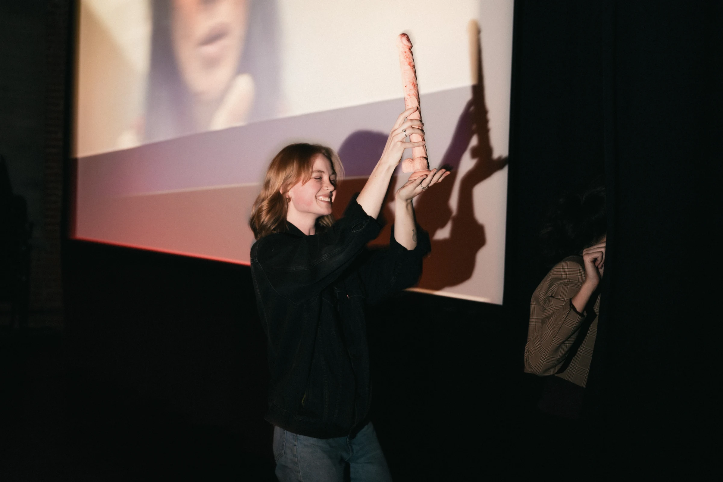 Person holding a tall, textured pink object up in front of a projector screen, with another person partially visible behind a curtain.