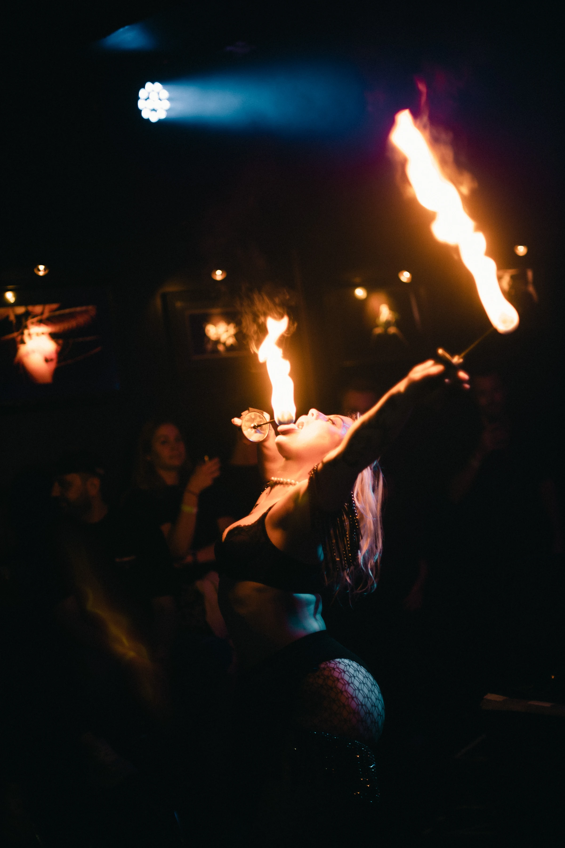 A performer in a dimly lit room breathes fire, holding two flaming torches. Audience members watch in awe, illuminated by warm light, creating a dramatic tone.