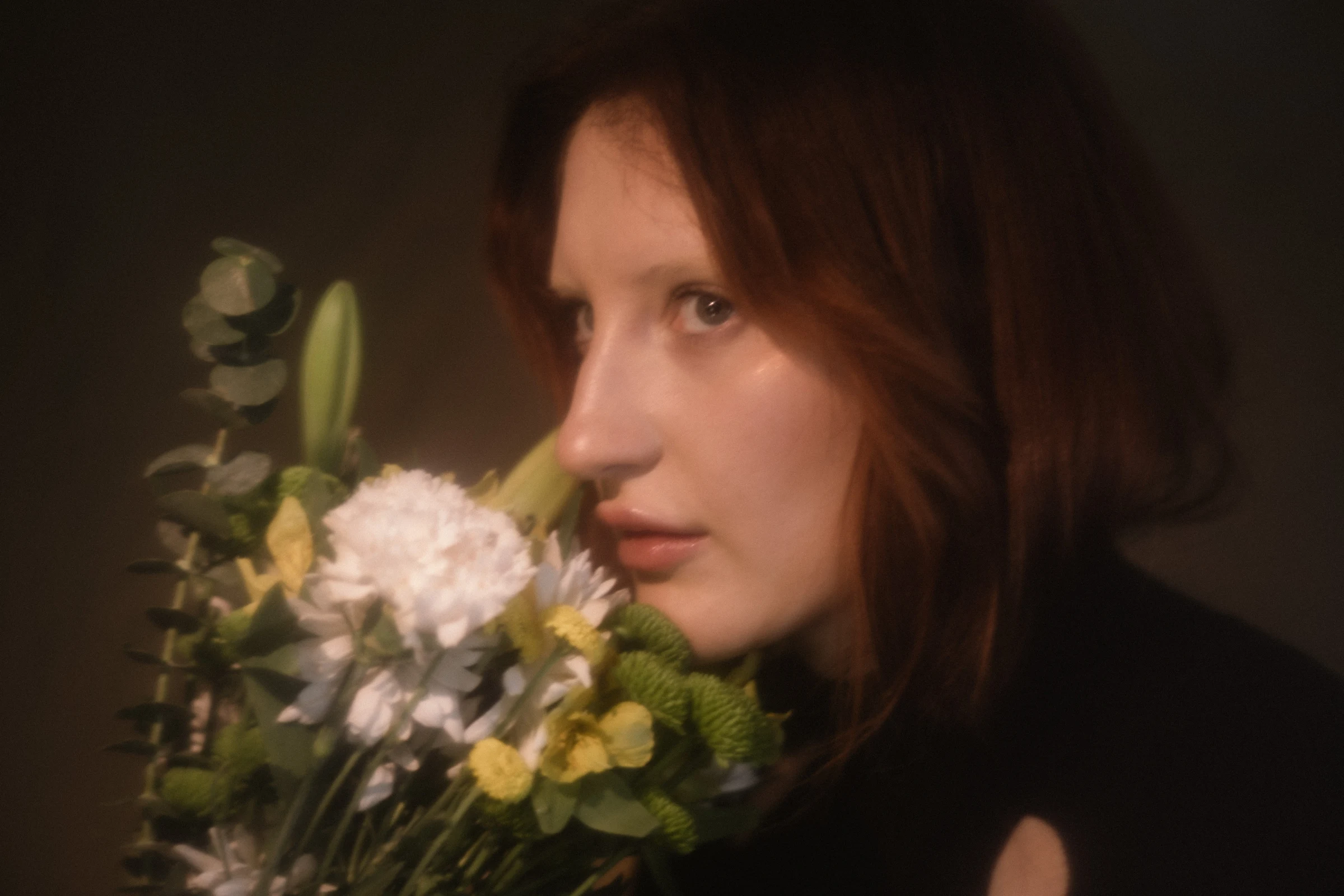 A woman with auburn hair gazes thoughtfully, holding a bouquet of white, yellow, and green flowers. The soft focus creates a dreamy, serene atmosphere.
