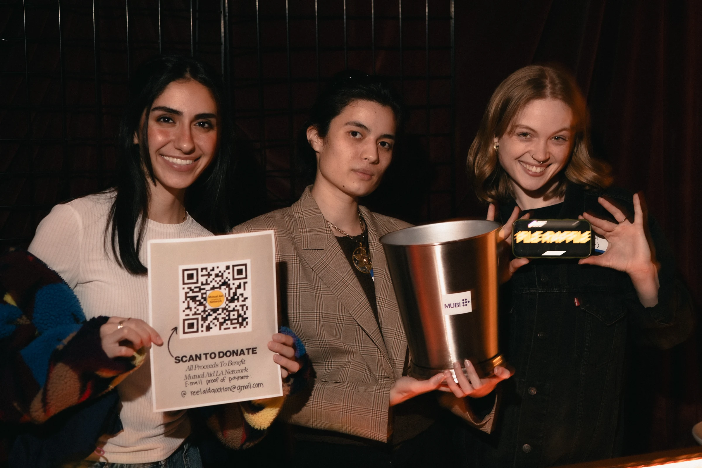 Three people smiling indoors. One holds a donation QR code sign, another has a metal bucket, and the third displays a neon "VEGANIC" sign. Warm, friendly atmosphere.
