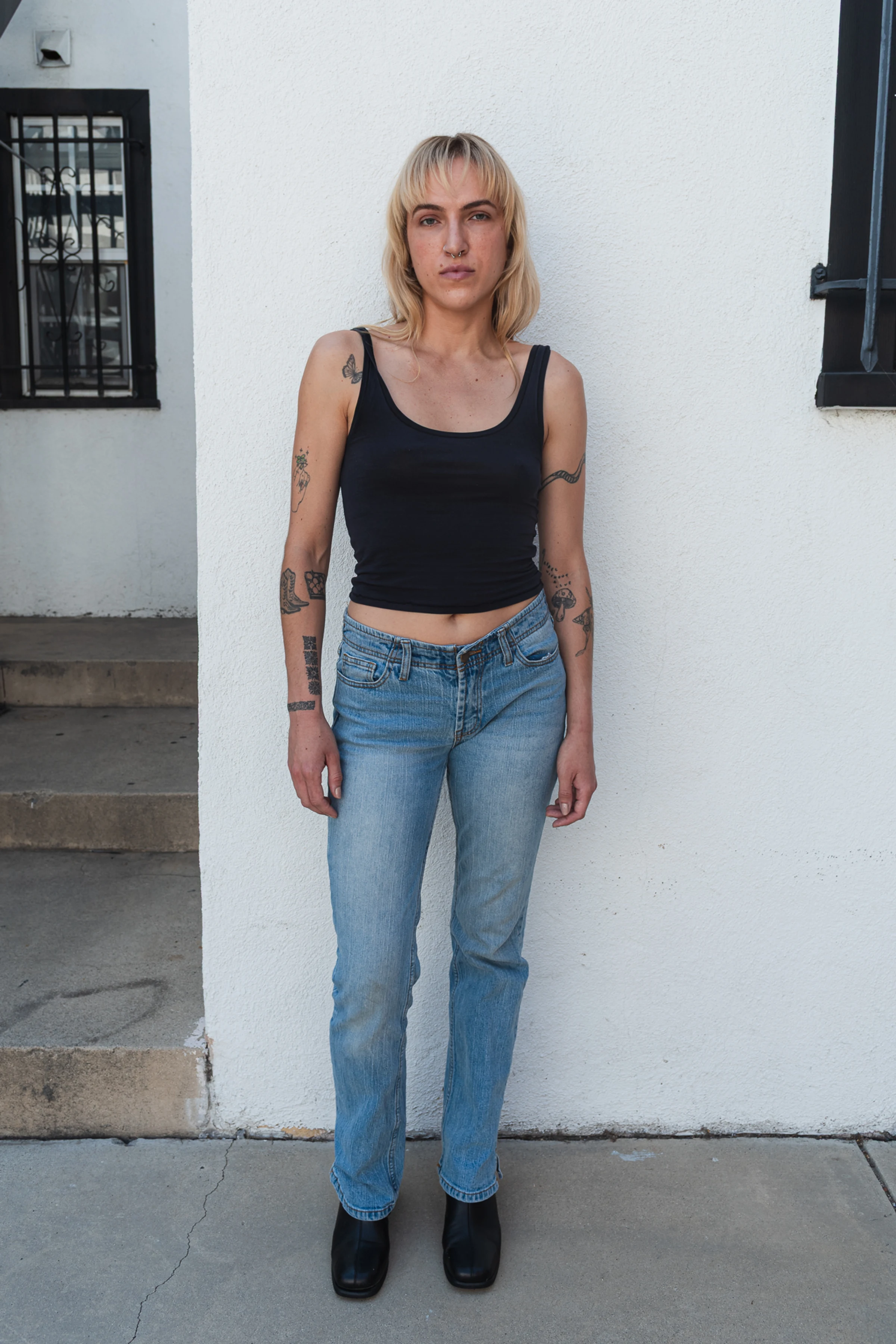 Person with tattoos stands against a white wall, wearing a black tank top and blue jeans. The setting is casual and urban with a neutral expression.