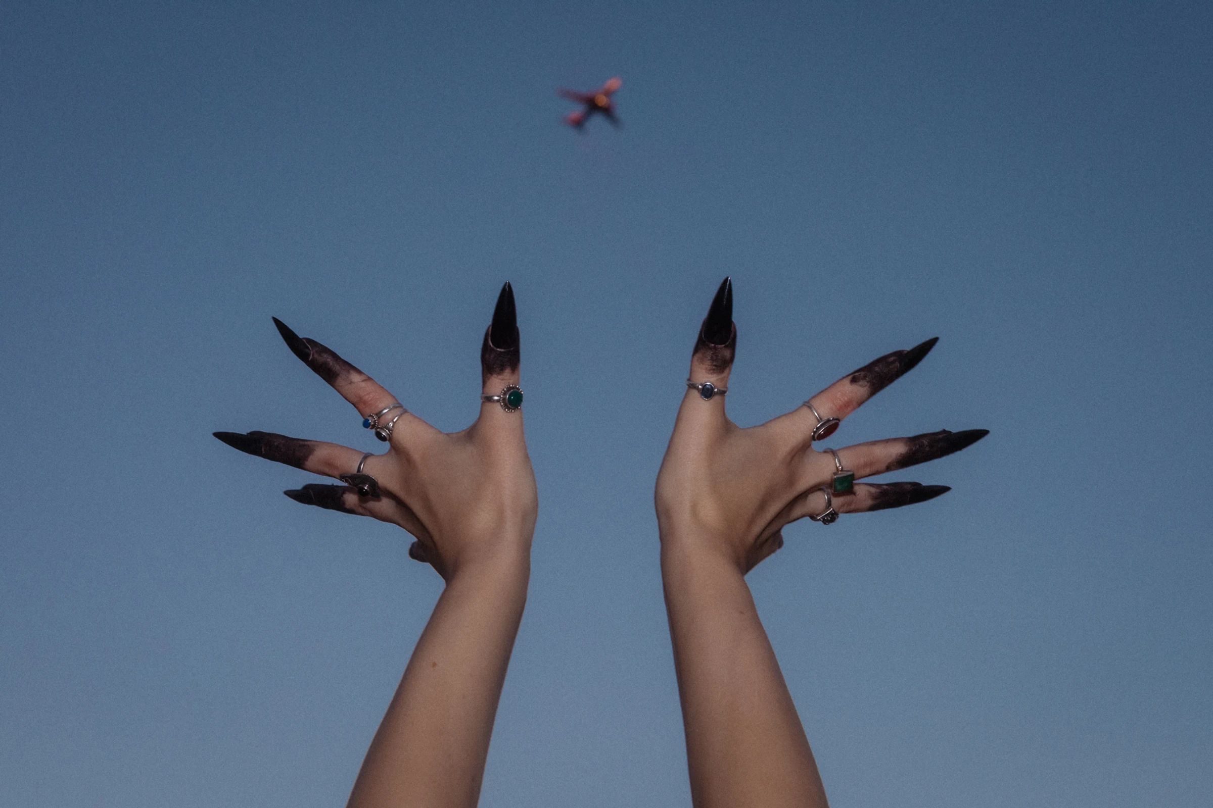 Two hands with long black nails, adorned with rings, reaching up against a clear blue sky with a small red airplane in the distance.