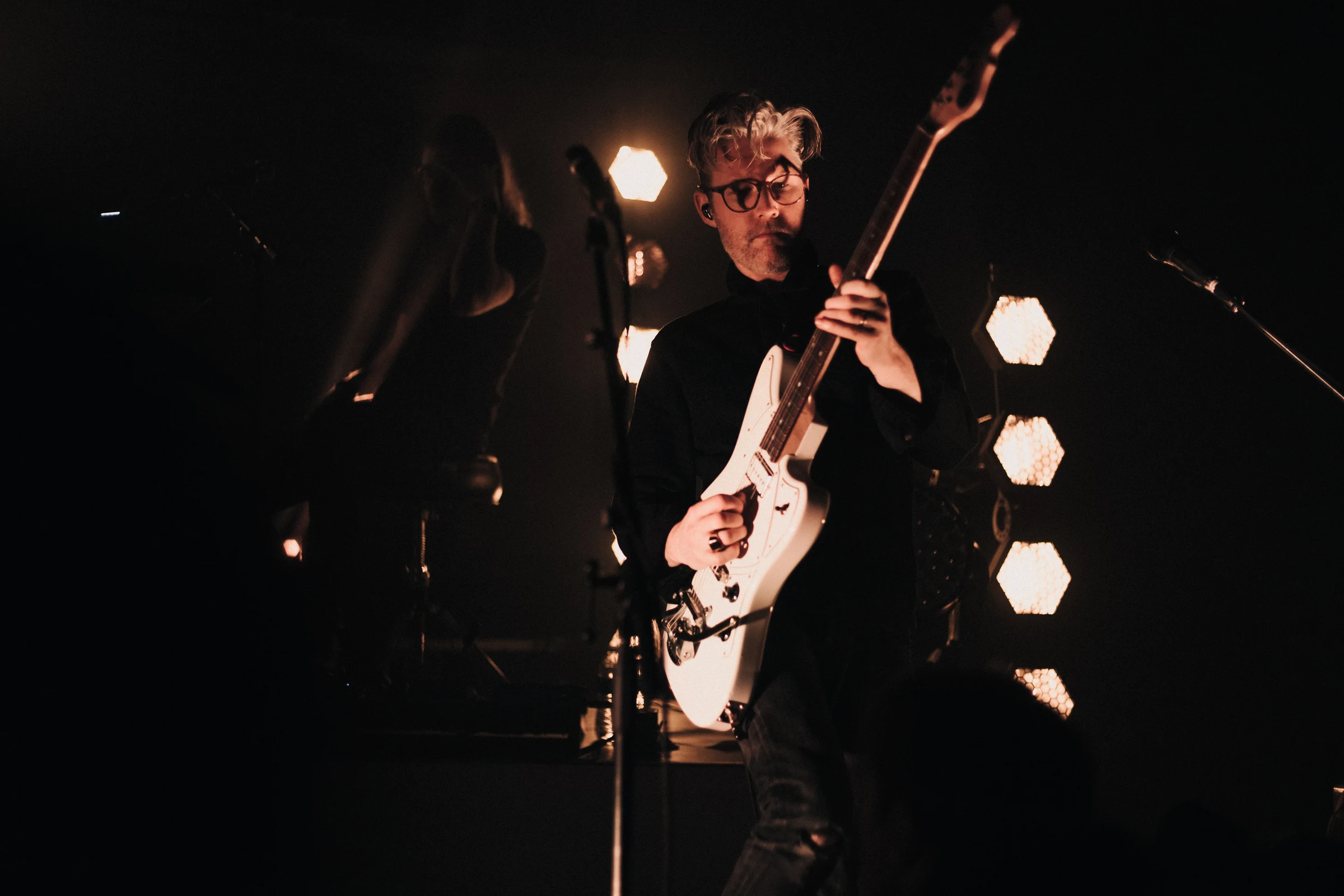 A guitarist wearing glasses performs passionately on stage, holding a white electric guitar. The dimly lit setting features hexagonal lights in the background.