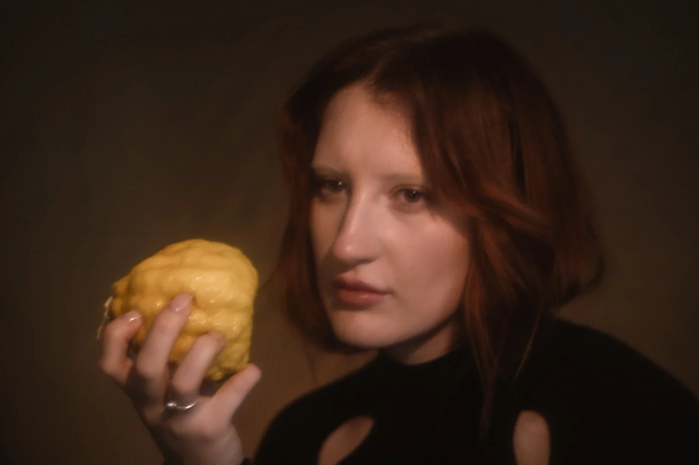 A woman with auburn hair holds a large, textured yellow fruit, possibly a citron. Her expression is contemplative, set against a soft, moody background.