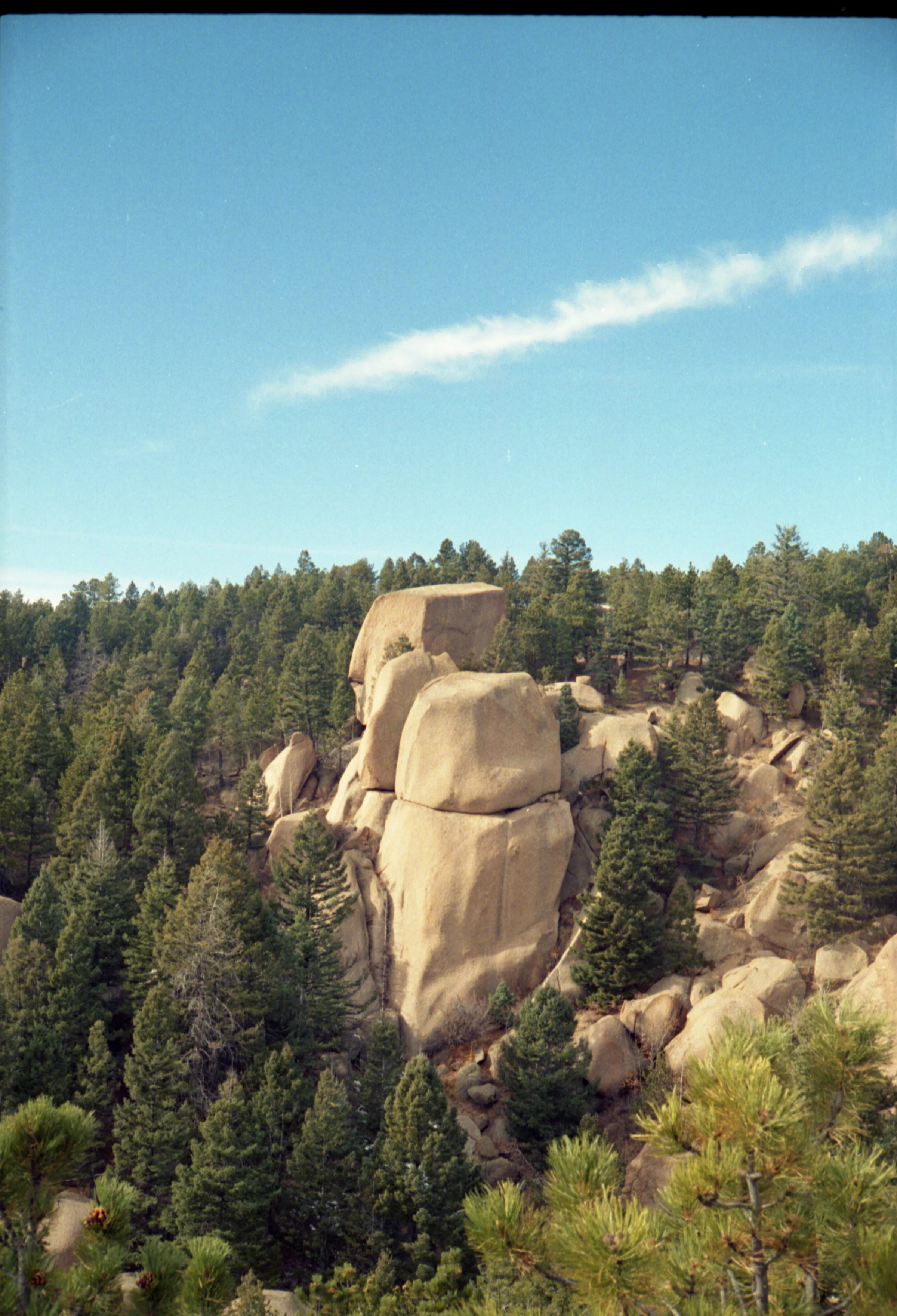 Rocky hillside with large boulders surrounded by dense evergreen forest. A clear blue sky with a streak of wispy clouds creates a serene atmosphere.