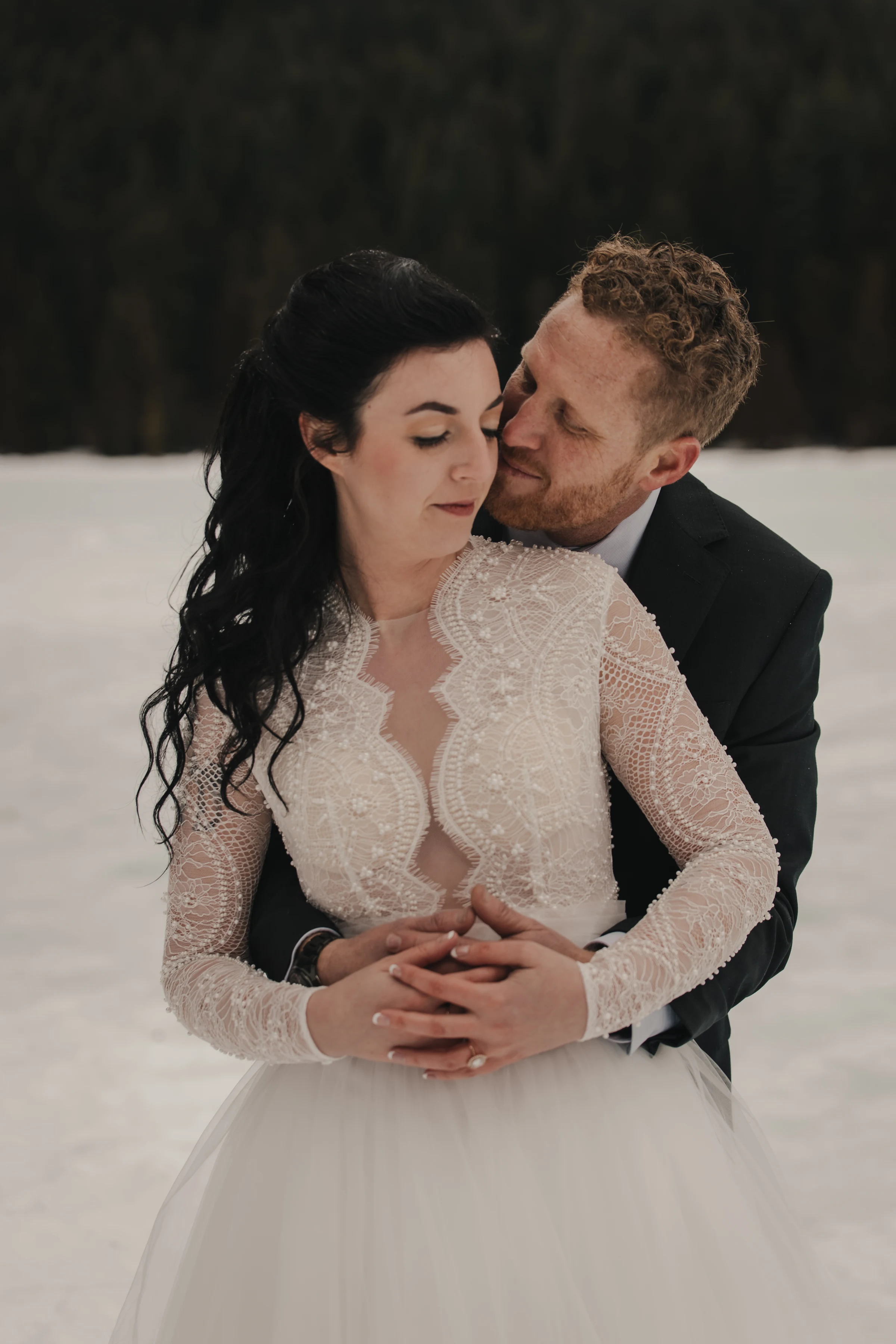 A bride in an intricate lace gown and a groom in a suit embrace tenderly on a snowy landscape, conveying love and warmth amidst a serene winter backdrop.
