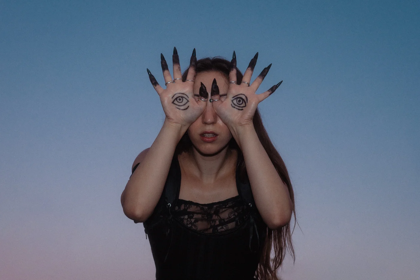 A woman poses against a twilight sky, holding her hands over her face with eyes drawn on the palms. She wears long, dark nails, conveying a surreal and mysterious tone.