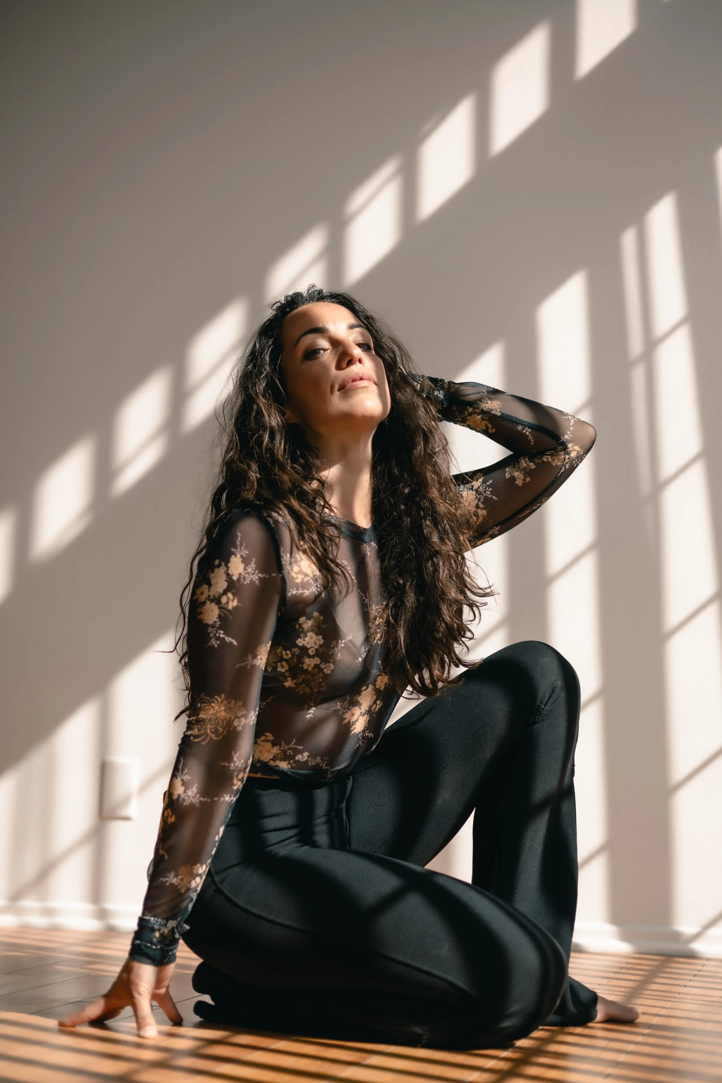 Woman kneels on a sunlit wooden floor, wearing a floral sheer top and black pants, hair loose. Shadows create a geometric pattern, conveying a serene mood.