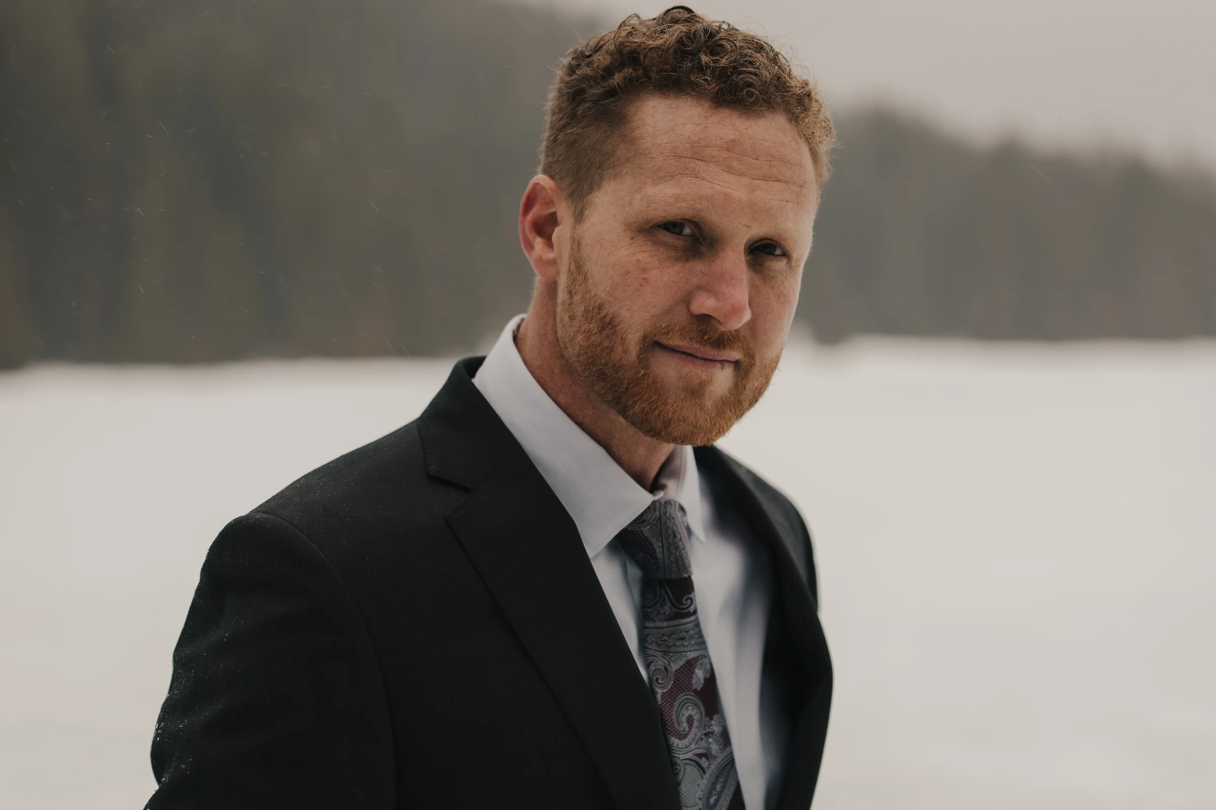 Man in a suit with a patterned tie stands outdoors, surrounded by a snowy landscape and blurred forest. He appears contemplative and confident.