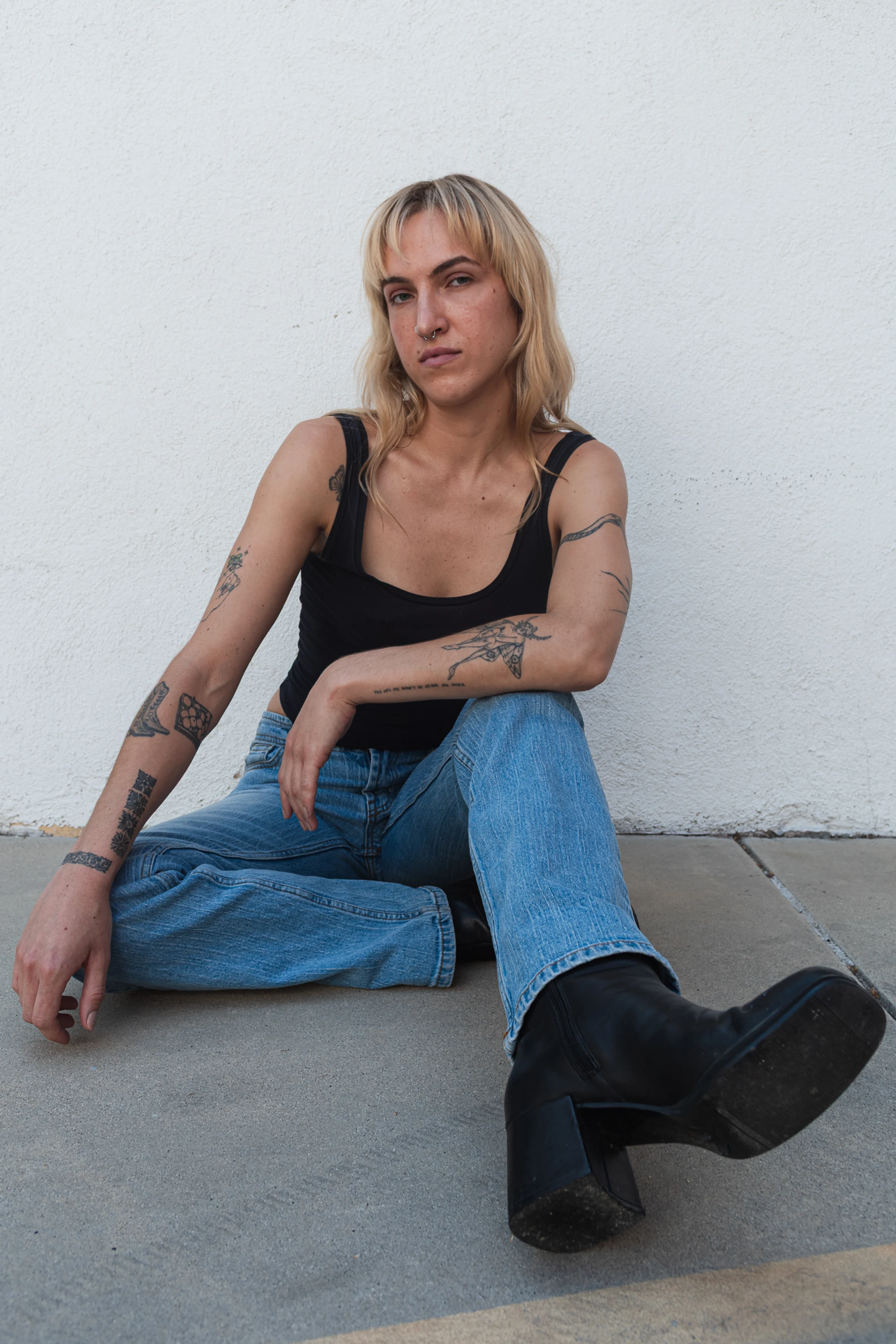 A person with long, blonde hair sitting against a white wall. They wear a black tank top, blue jeans, and black boots, with tattoos on their arms, conveying a relaxed and confident vibe.