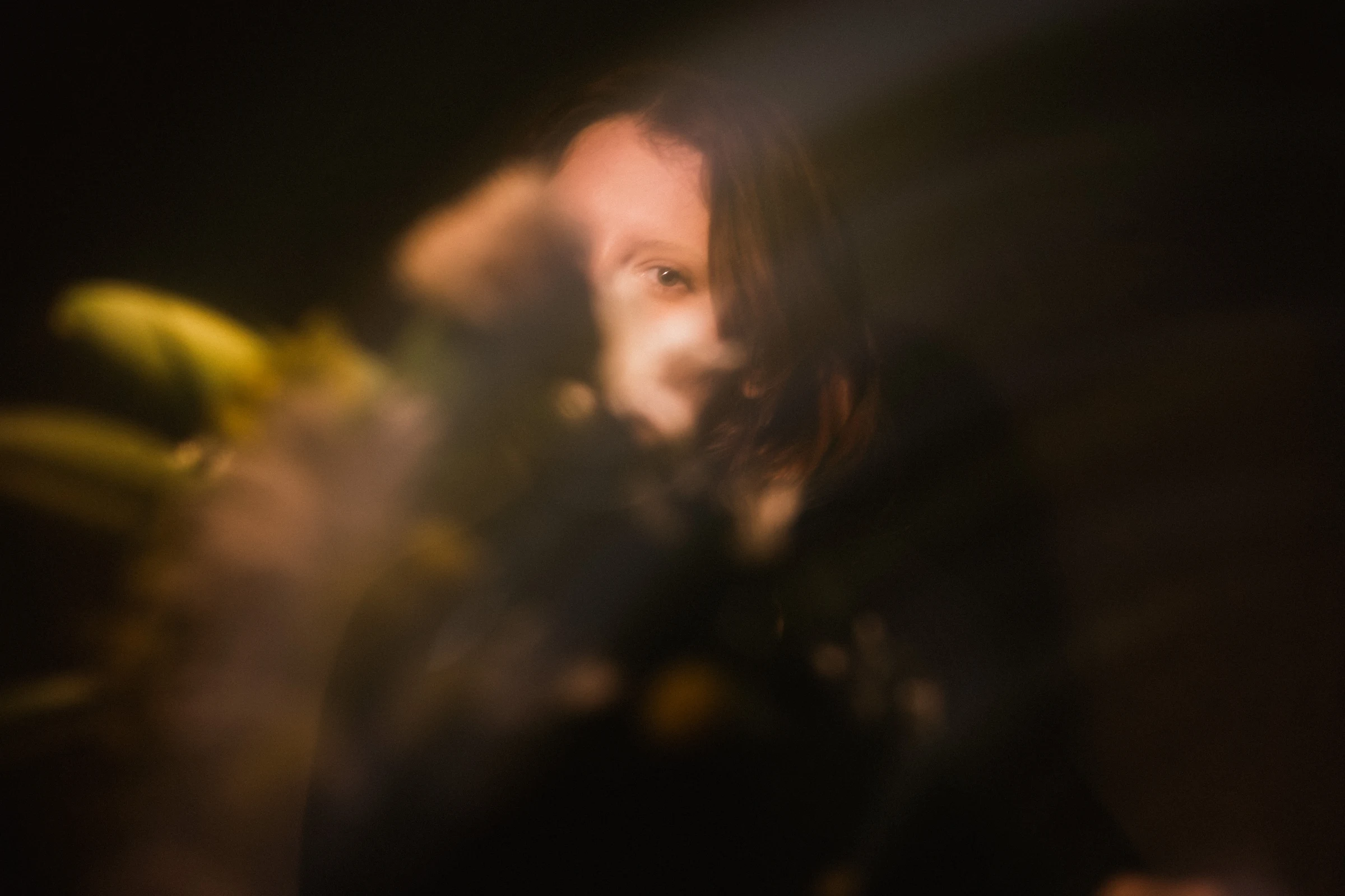 A blurred, moody portrait shows a person partially obscured by soft focus leaves, with one eye visible. Warm, dim lighting adds a mysterious feeling.