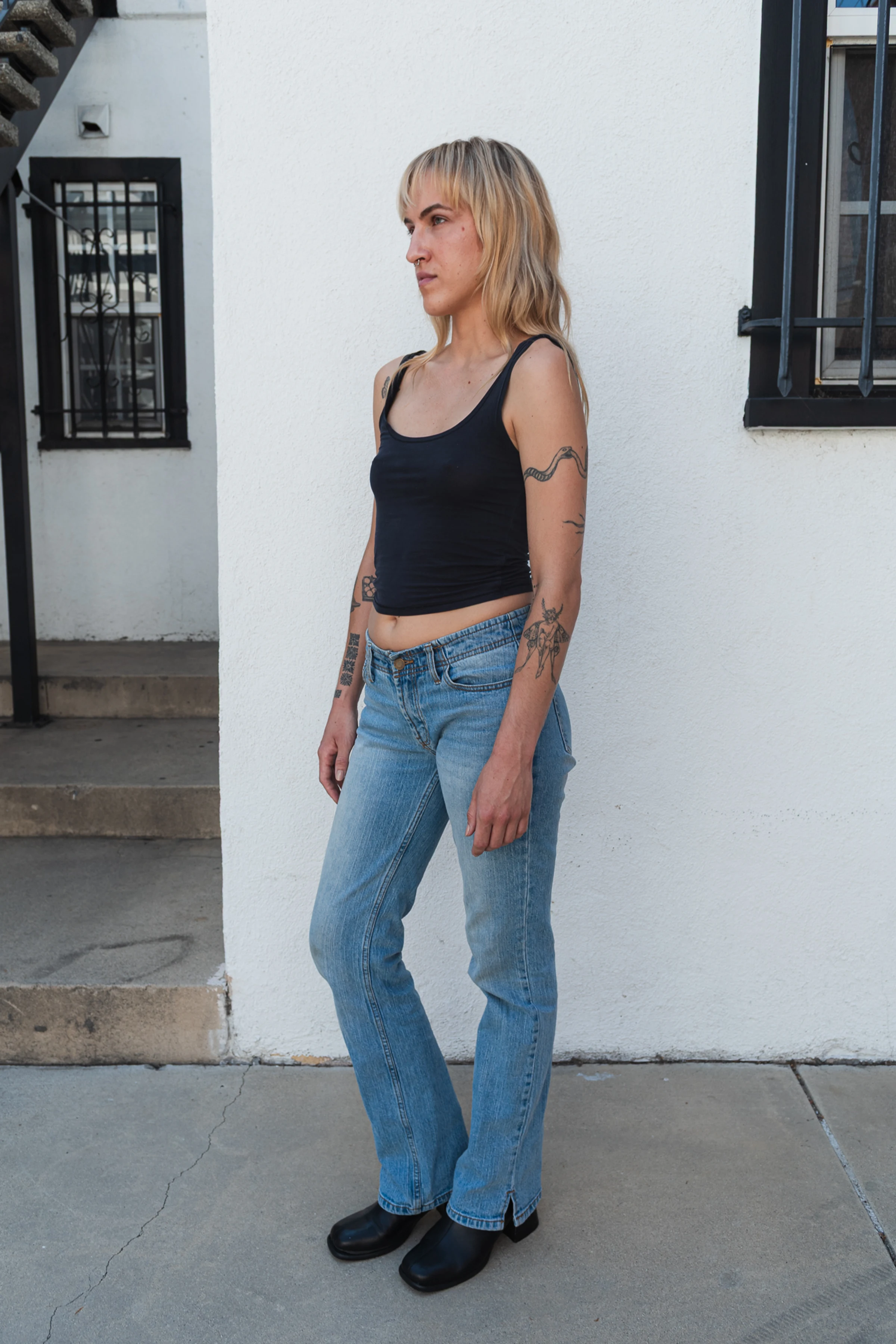 A person with long blond hair stands in profile against a white wall. They wear a black tank top, blue jeans, and black boots. Tattoos are visible on their arms.