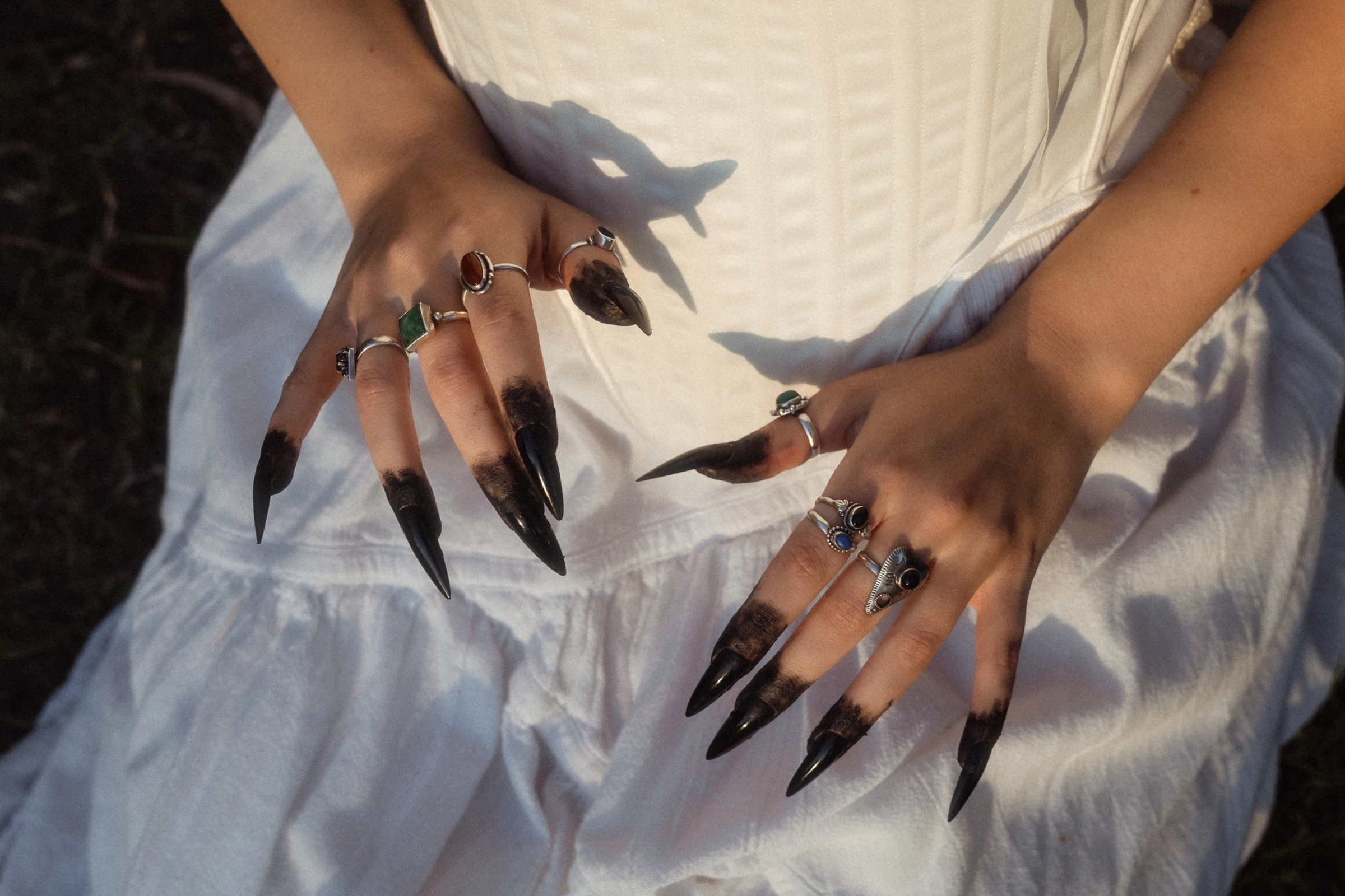 Hands with long black pointed nails and multiple rings resting on a white dress.