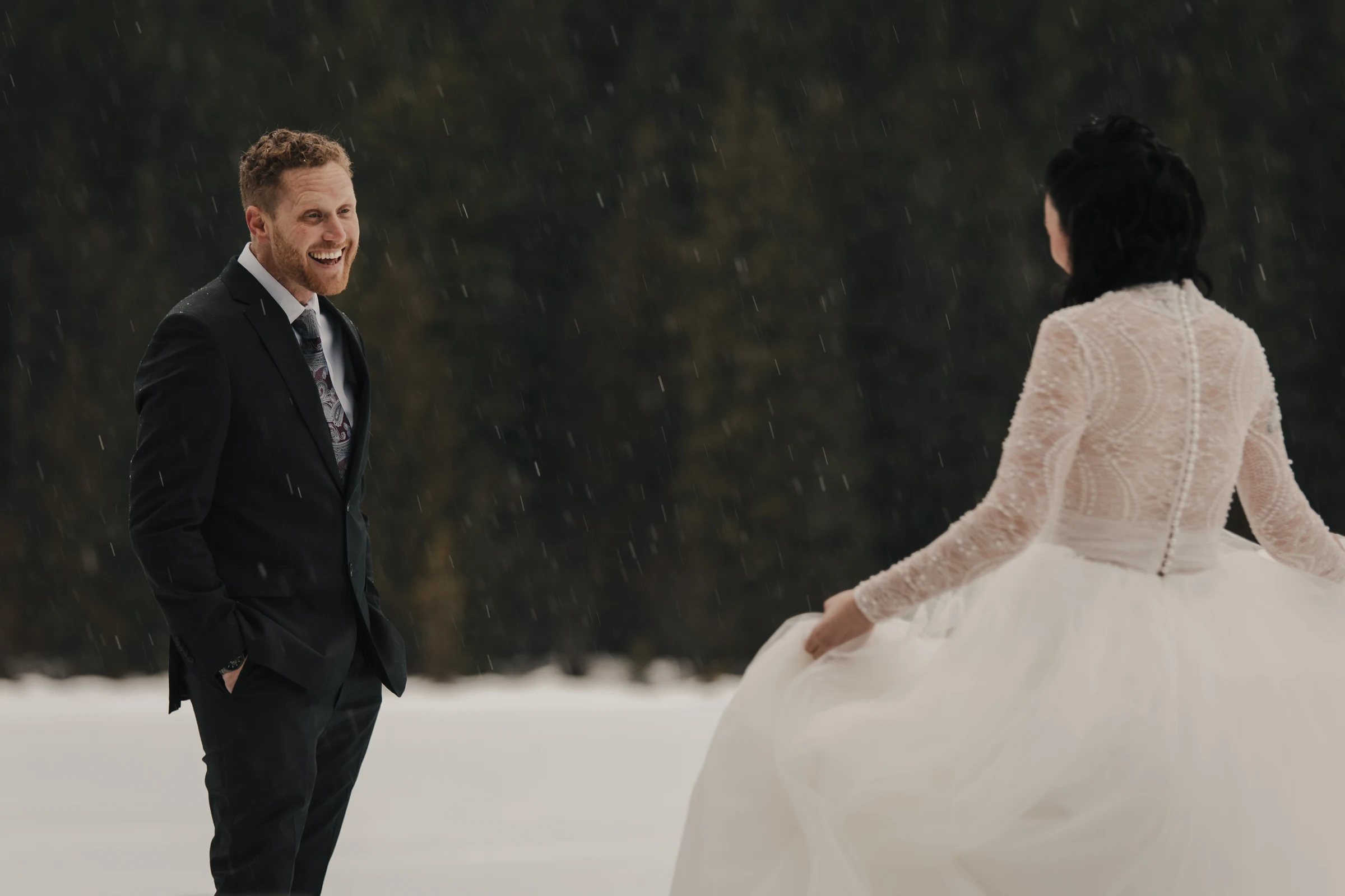 Bride in a white gown and groom in a black suit stand facing each other outdoors in falling snow.