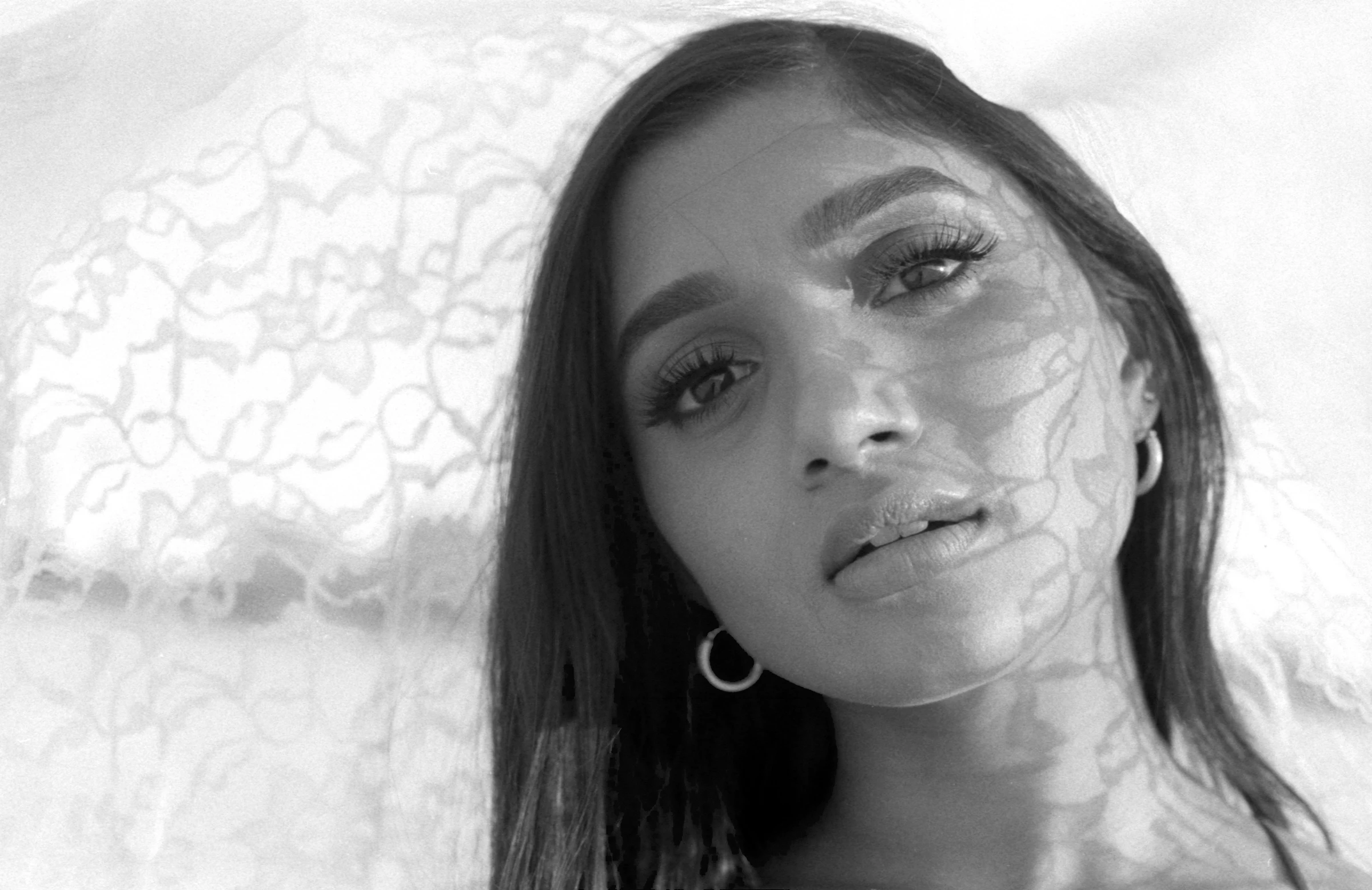 Black and white portrait of a woman with lace shadows on her face, gazing softly. Her long hair frames her expression, evoking a serene mood.