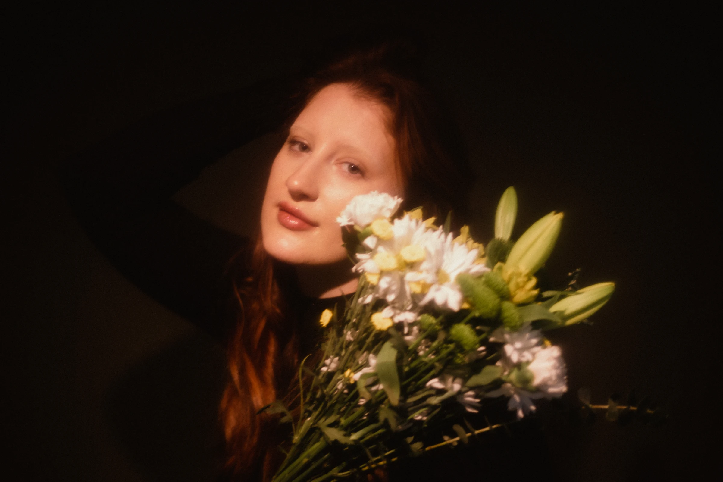 A woman with long, red hair poses in dim lighting, holding a bouquet of white flowers and green leaves. The mood is soft and serene.