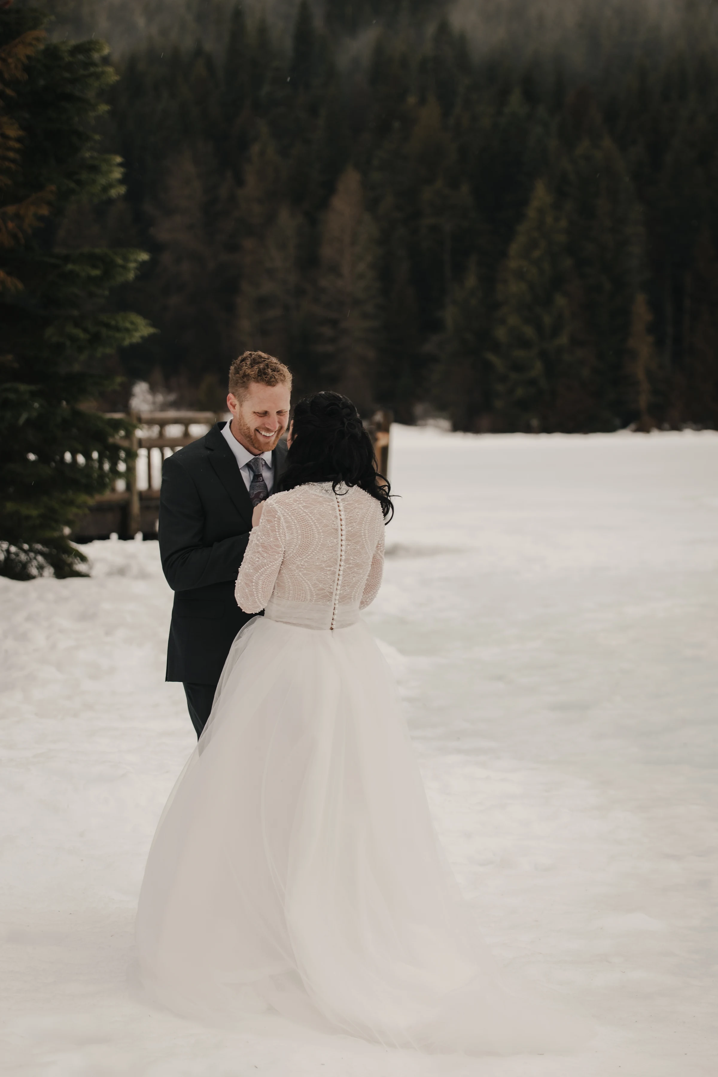 A couple stands on snow, embracing in front of a forest. The bride wears a white gown; the groom a dark suit, both smiling warmly, conveying joy.