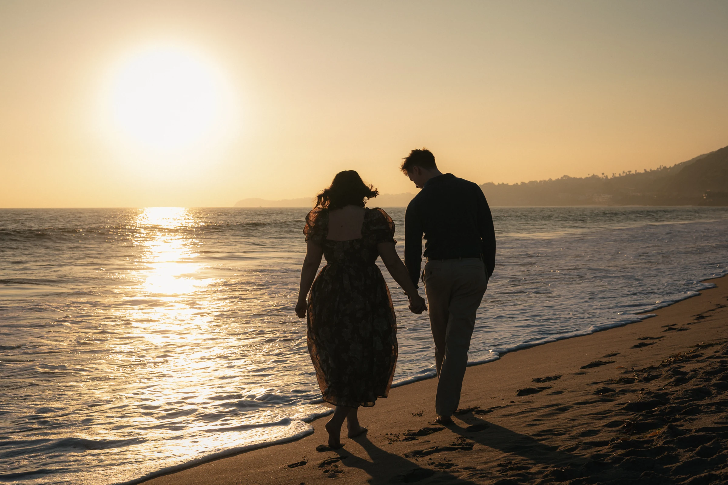 A couple holding hands walks along a sandy beach at sunset. The ocean reflects the golden sun, creating a serene and romantic atmosphere.