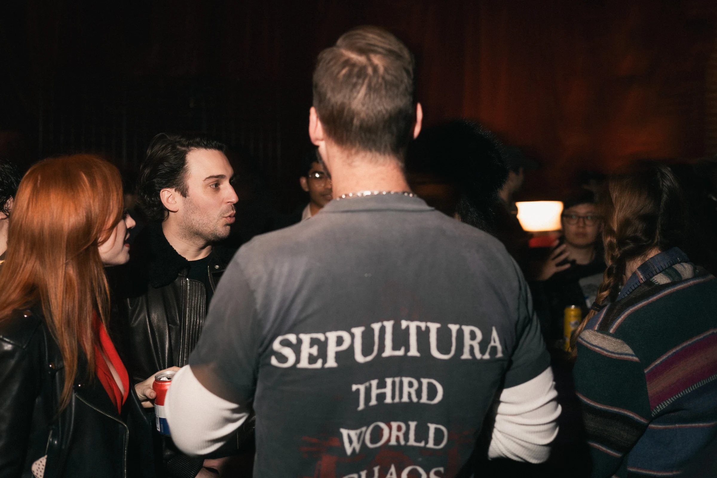 A group of people socializing at a dimly lit gathering. One person wears a "Sepultura Third World Chaos" shirt. The mood is lively and casual.