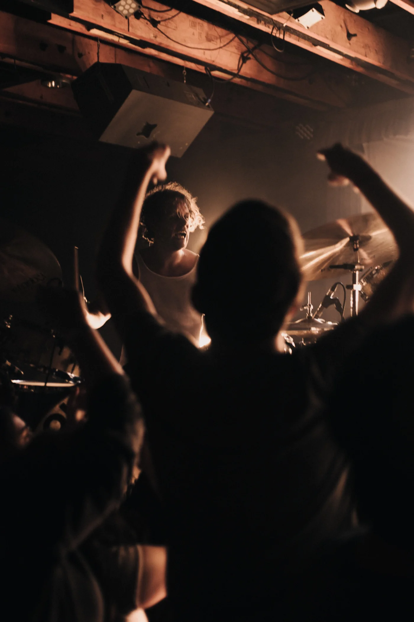 Drummer plays energetically on stage, surrounded by an enthusiastic crowd with raised arms, in a dimly lit, intimate concert setting.