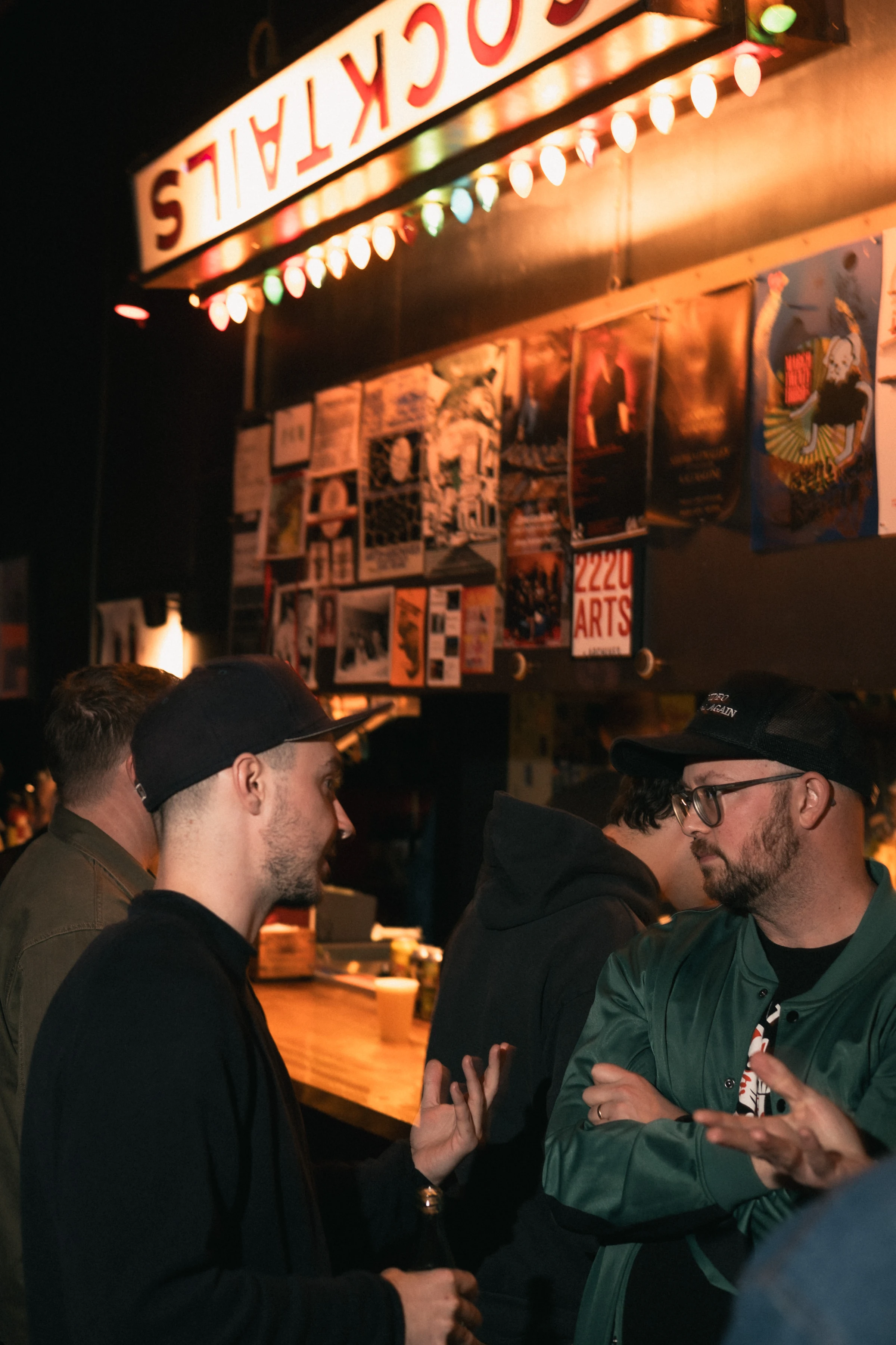 Two men converse in a dimly lit bar beneath a neon "Cocktails" sign. The background features a wall adorned with colorful posters. Mood is lively and relaxed.
