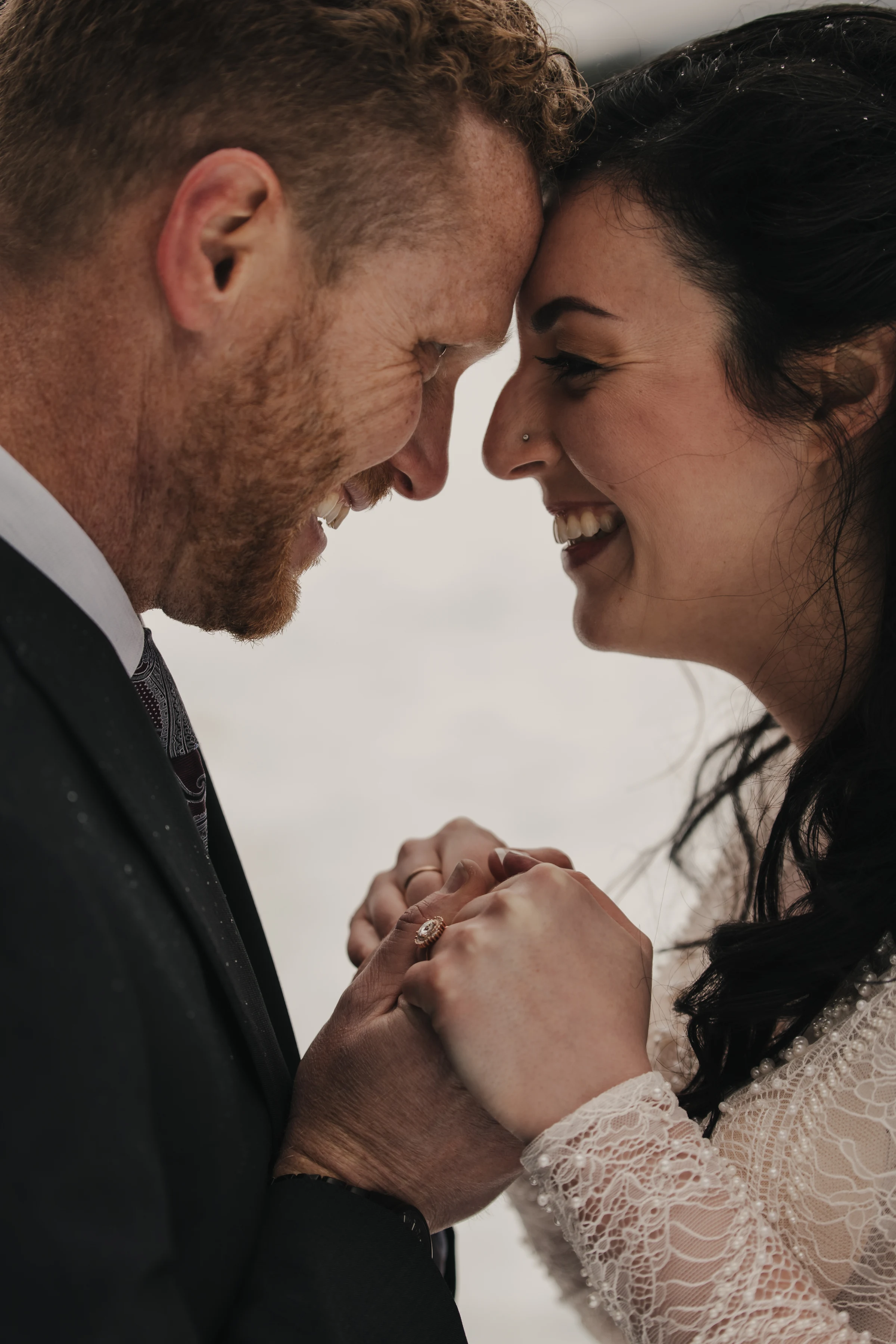 A joyful couple shares an intimate moment, touching foreheads and holding hands. They are smiling warmly, conveying love and happiness on a snowy day.