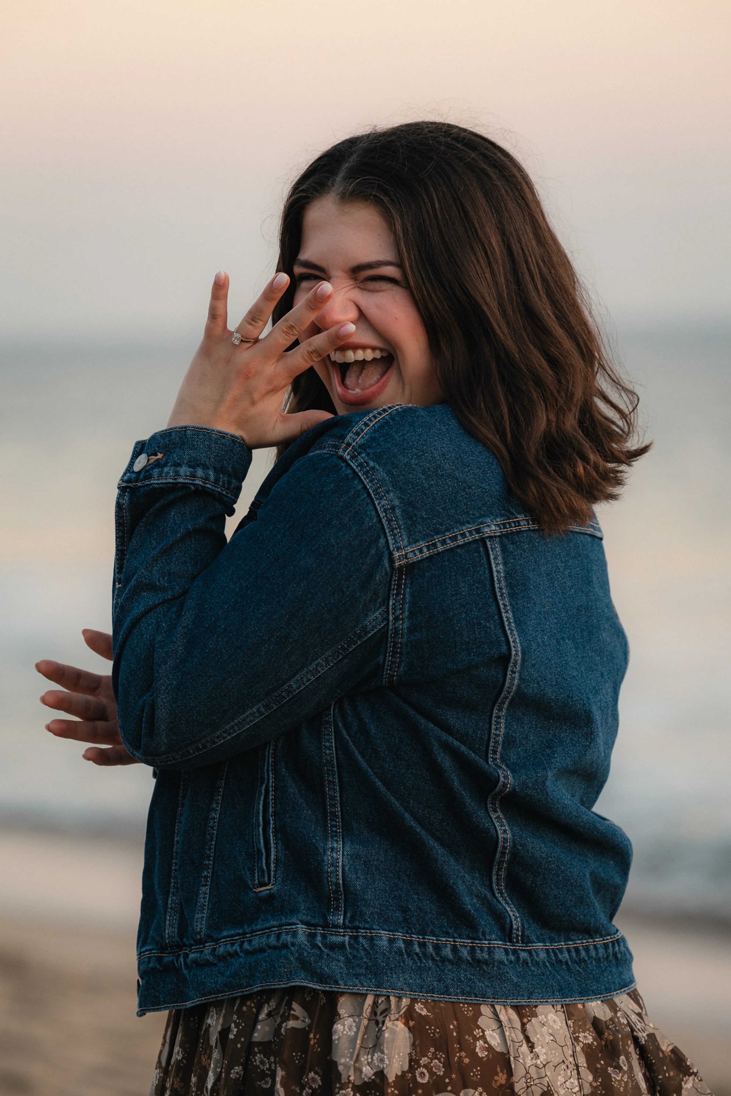 Young woman in denim jacket laughing joyfully with her hand playfully touching her face, standing on a beach at sunset, conveying happiness.
