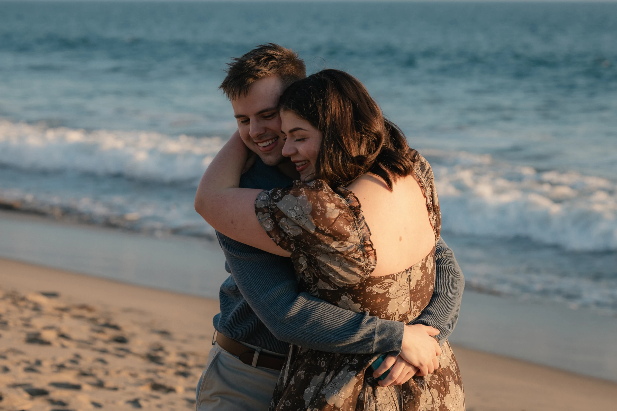 A couple embraces warmly on a beach, smiling with eyes closed. The ocean waves gently roll in the background, creating a serene atmosphere.