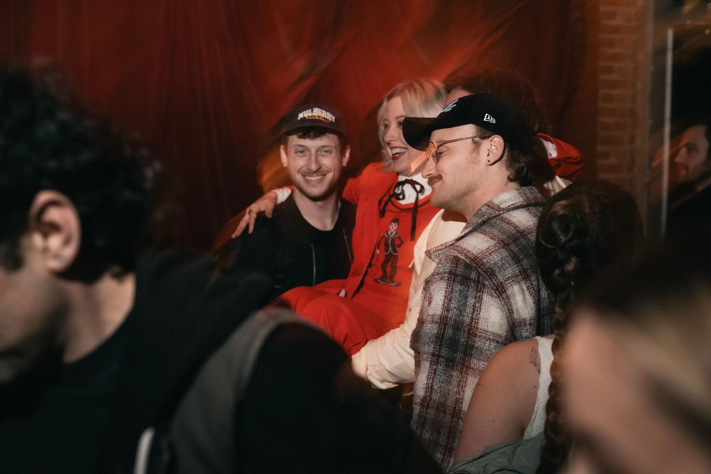 Group of friends enjoying a lively gathering. A woman in a red outfit is being playfully lifted, surrounded by smiling people in a dimly lit room.