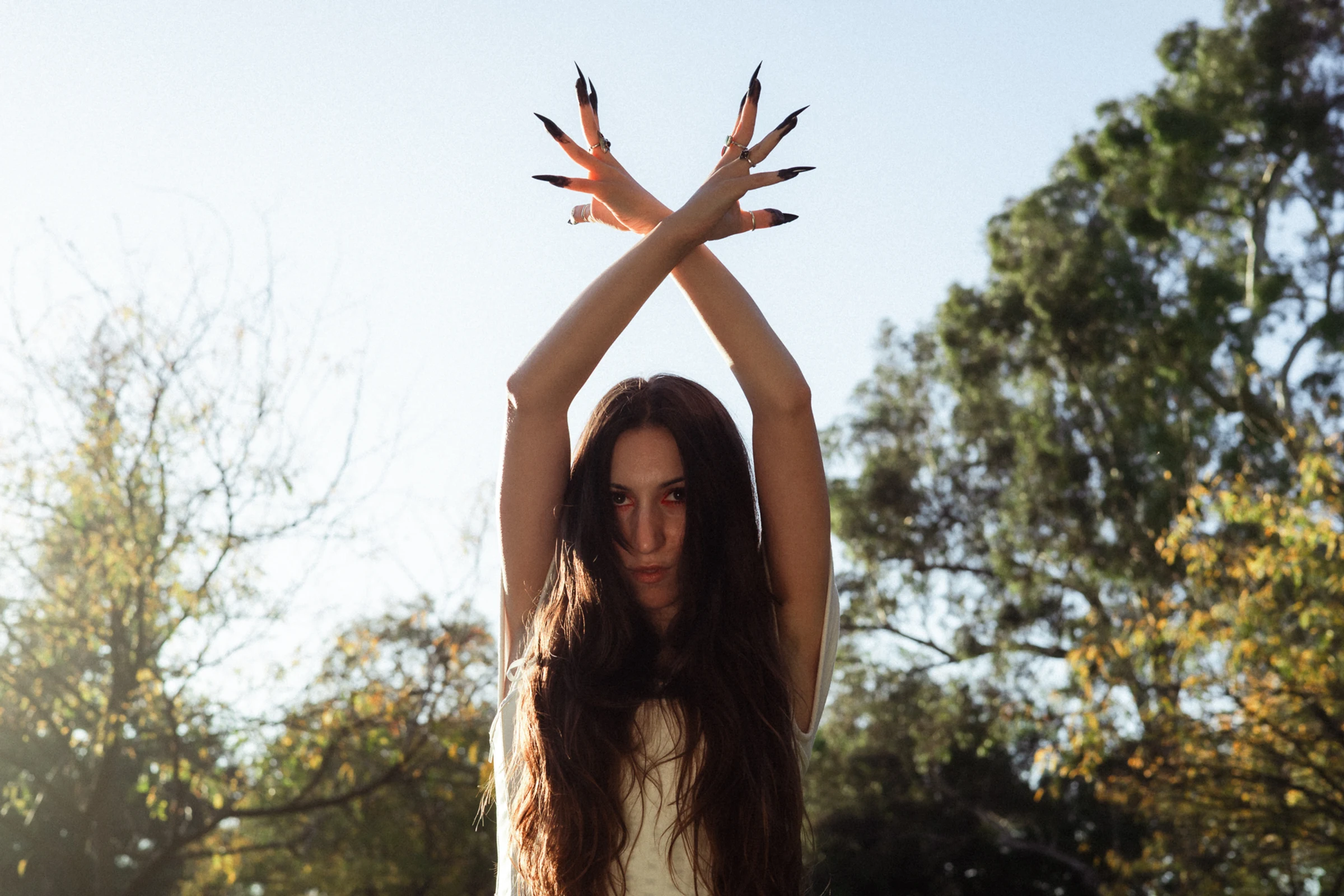 A woman with long hair stands outdoors, arms crossed above her head, fingers with long dark nails, evoking mystery. Trees are visible in the background.