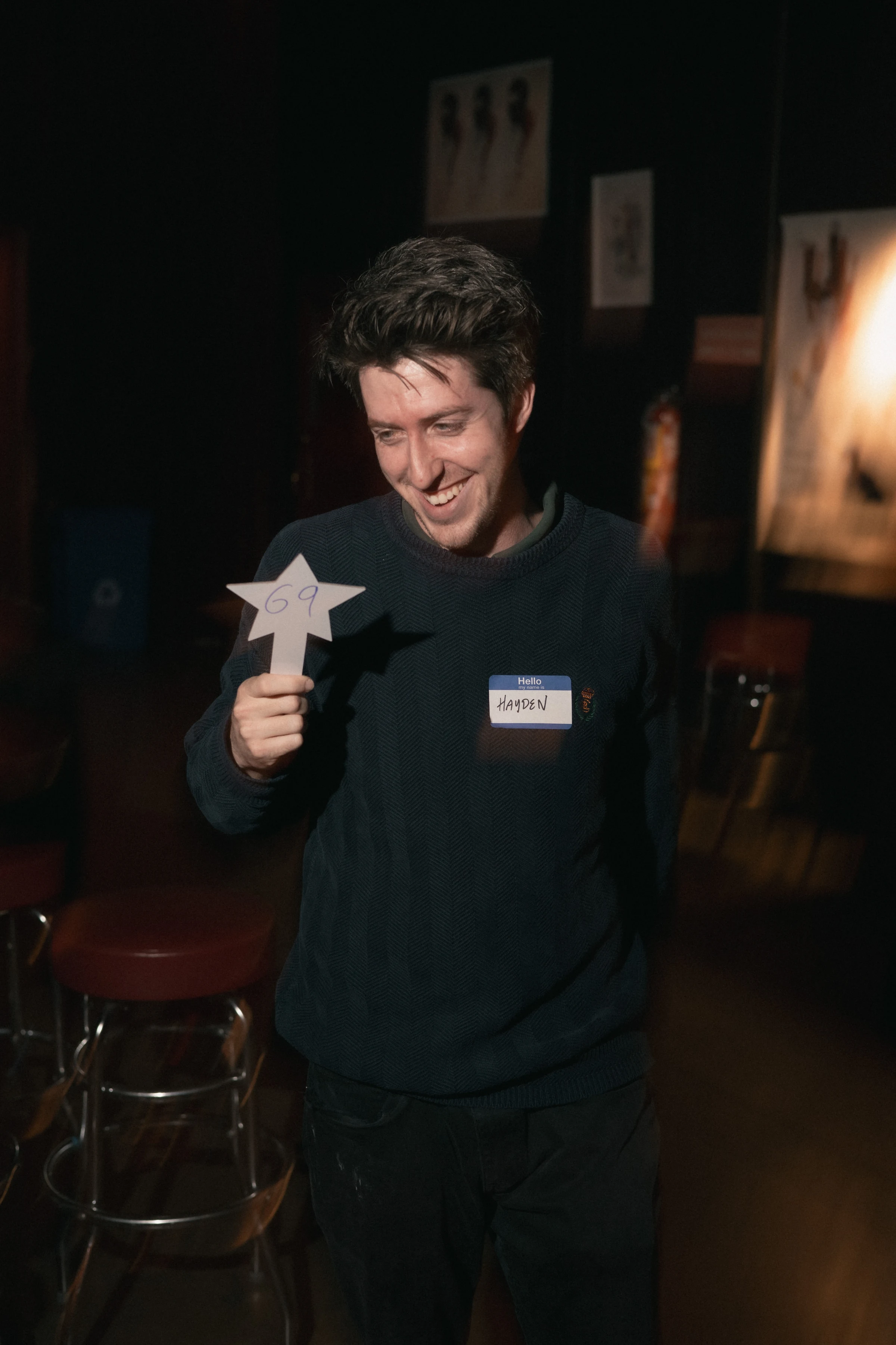 A man smiles while holding a paper star with "69" on it in a dimly lit room. He wears a dark sweater and a name tag. The mood is cheerful and informal.