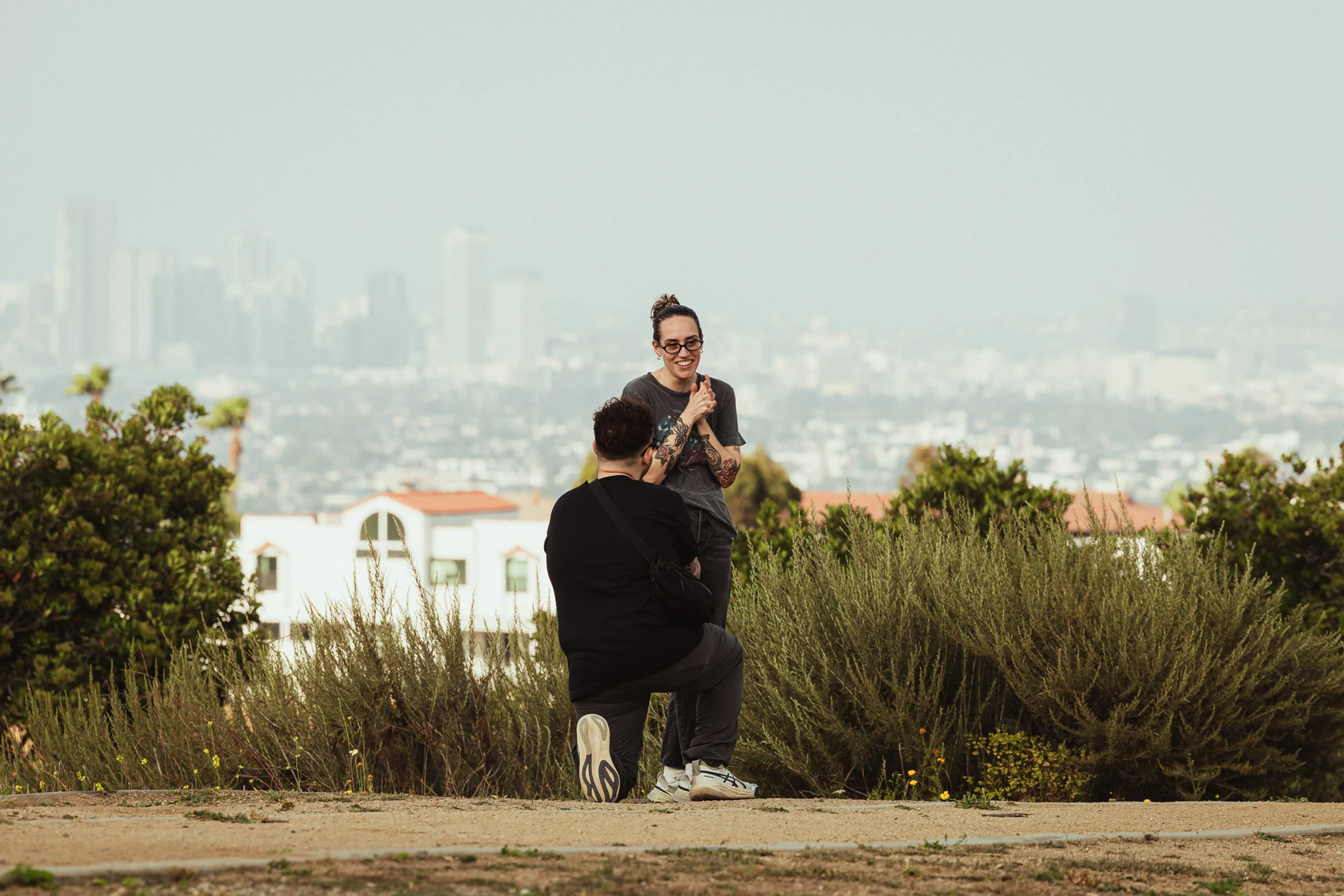 A person kneels, proposing to another who looks surprised and joyful. They stand on a hill with a cityscape in the misty background, surrounded by greenery.