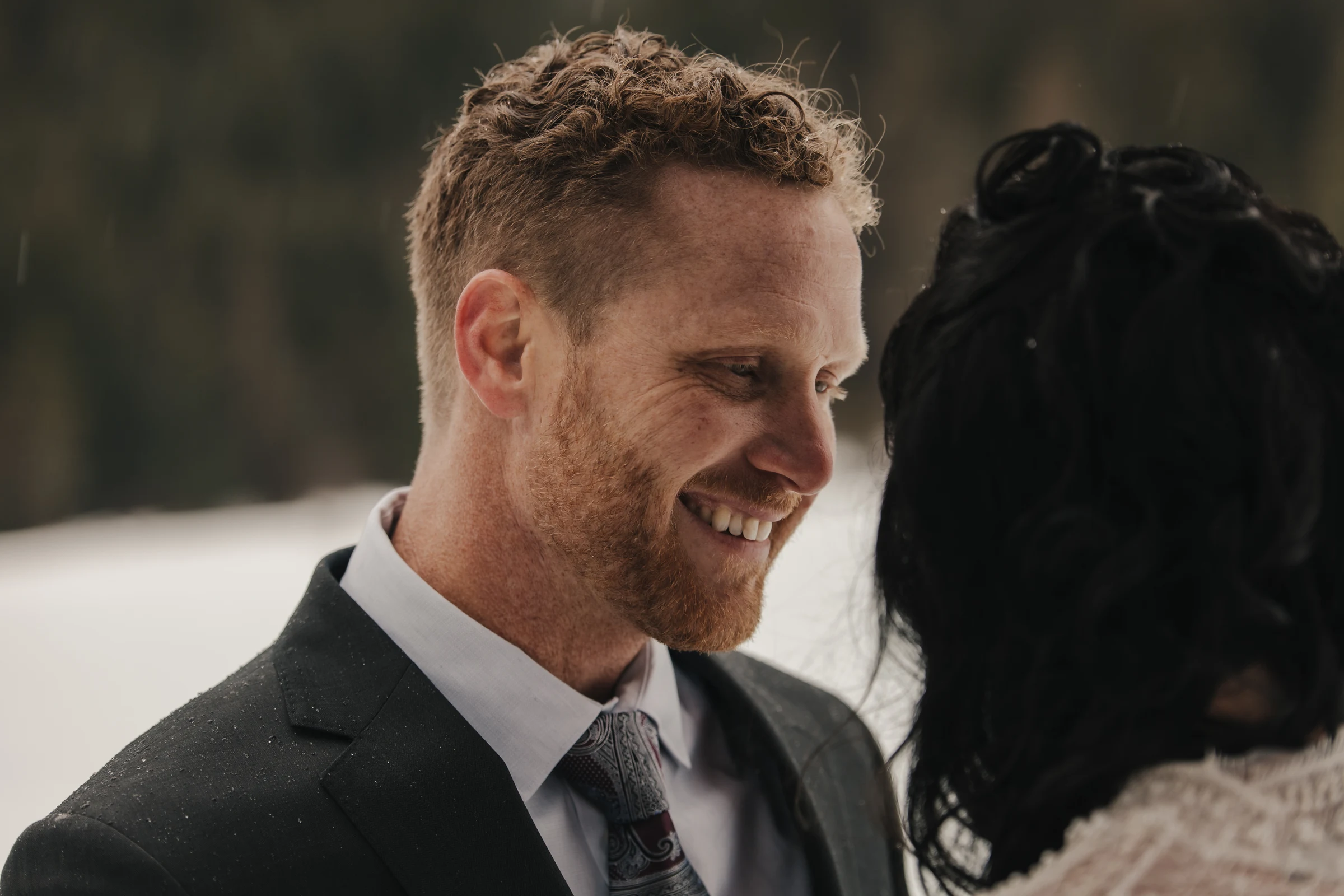 A man in a suit smiles warmly at a woman in a snowy outdoor setting. The mood is intimate and joyful, suggesting a romantic moment.
