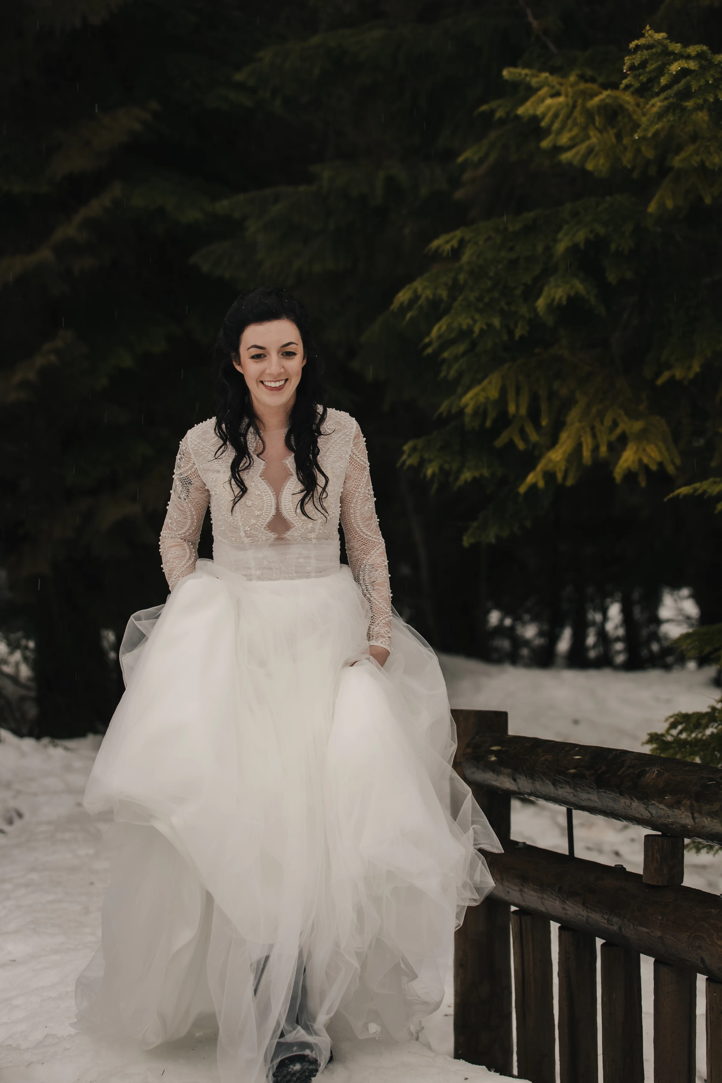 Bride in a lace wedding dress, smiling while walking through a snowy forest. The setting is serene, with green trees creating a natural backdrop.