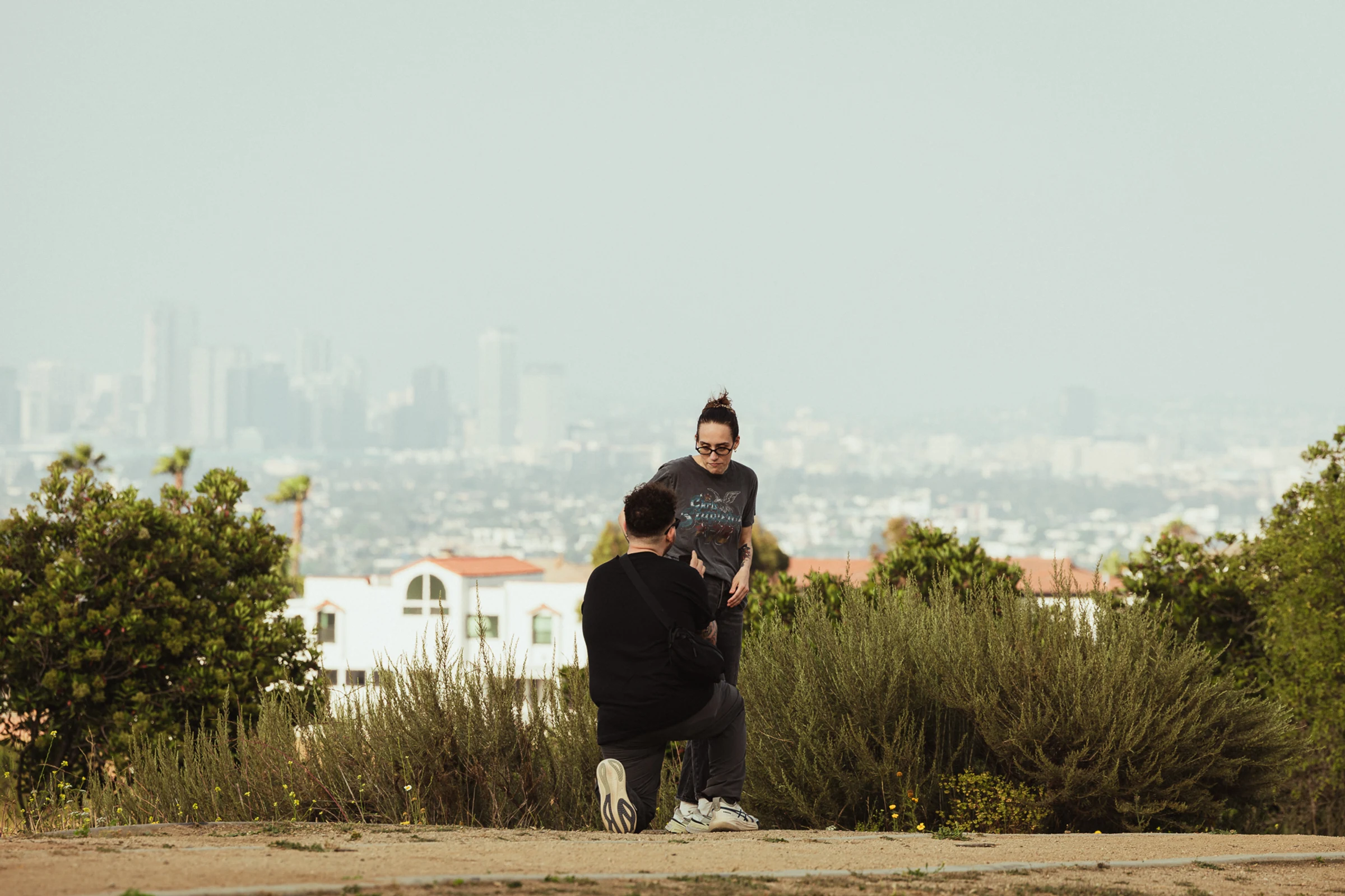 A person kneels, proposing to another in a park with a city skyline in the background. The scene conveys surprise and intimacy amidst lush greenery.