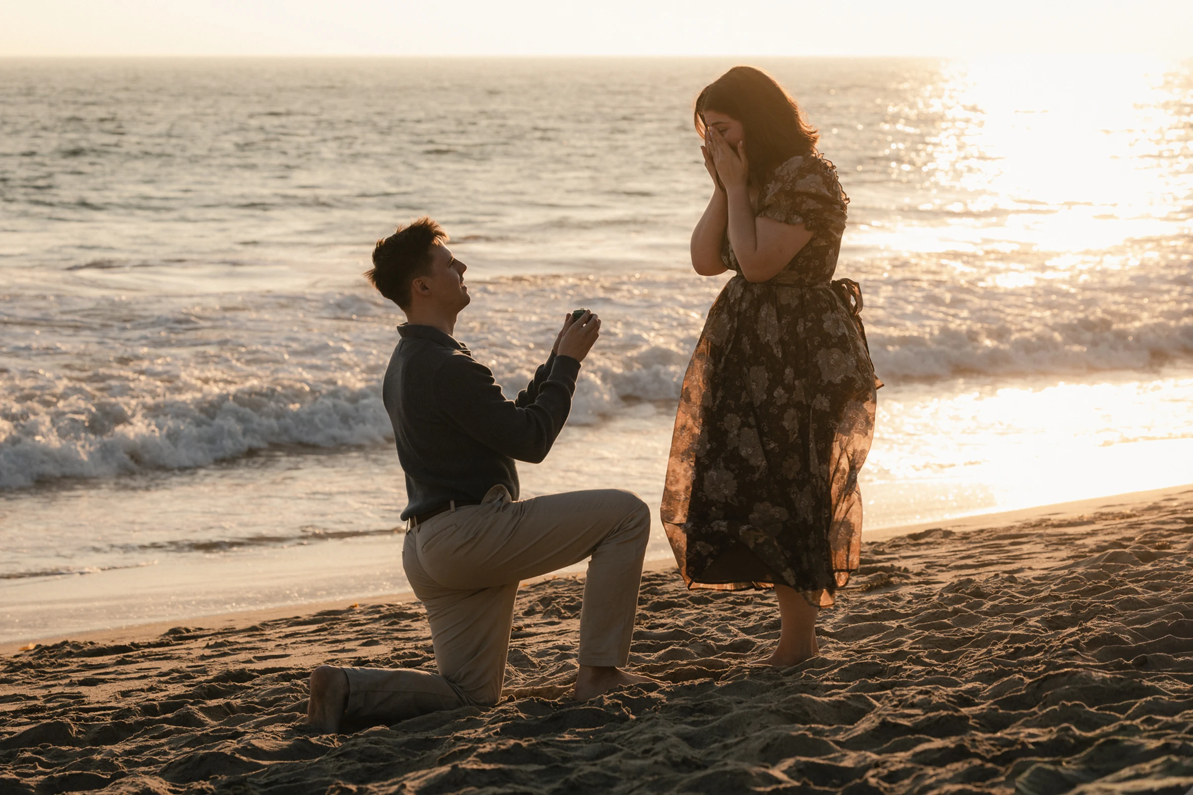 A man kneels on a beach at sunset, proposing to a surprised woman in a floral dress. The ocean waves and golden sunlight create a romantic atmosphere.