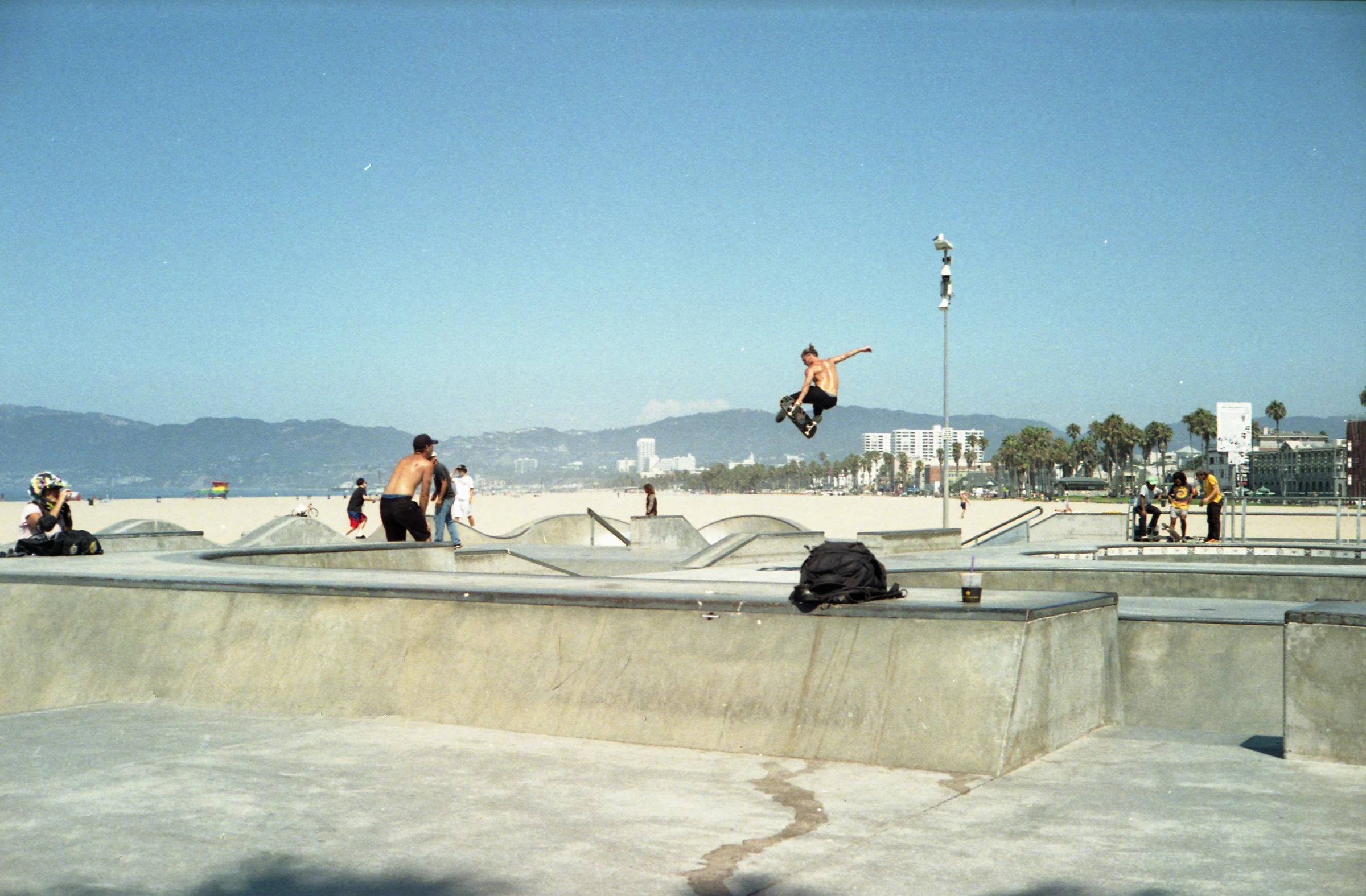 A shirtless skateboarder performs a high jump trick at a sunny beachfront skate park, with mountains and palm trees in the background. Energetic vibe.