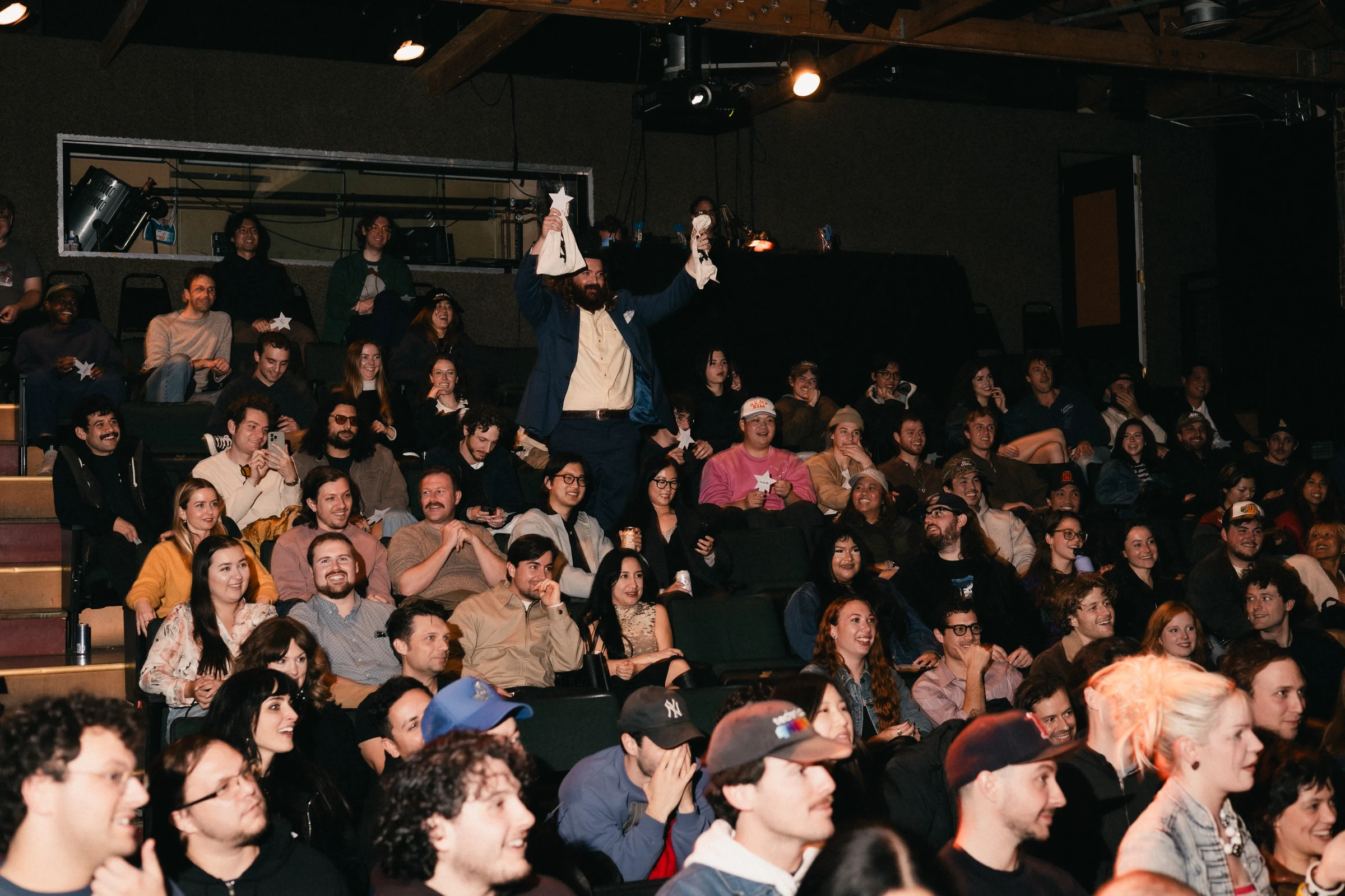 A lively audience fills a theater, showing excitement and enthusiasm. One person stands out, holding up white objects, creating an energetic atmosphere.