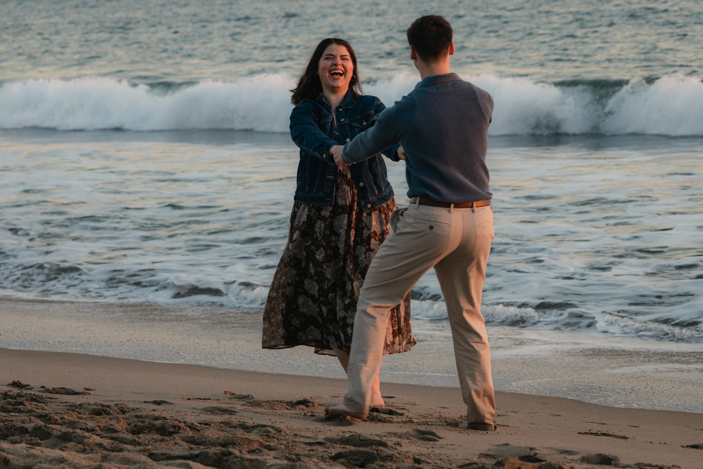 A couple joyfully dances on a sandy beach at sunset, with waves gently crashing in the background. The scene conveys happiness and warmth.