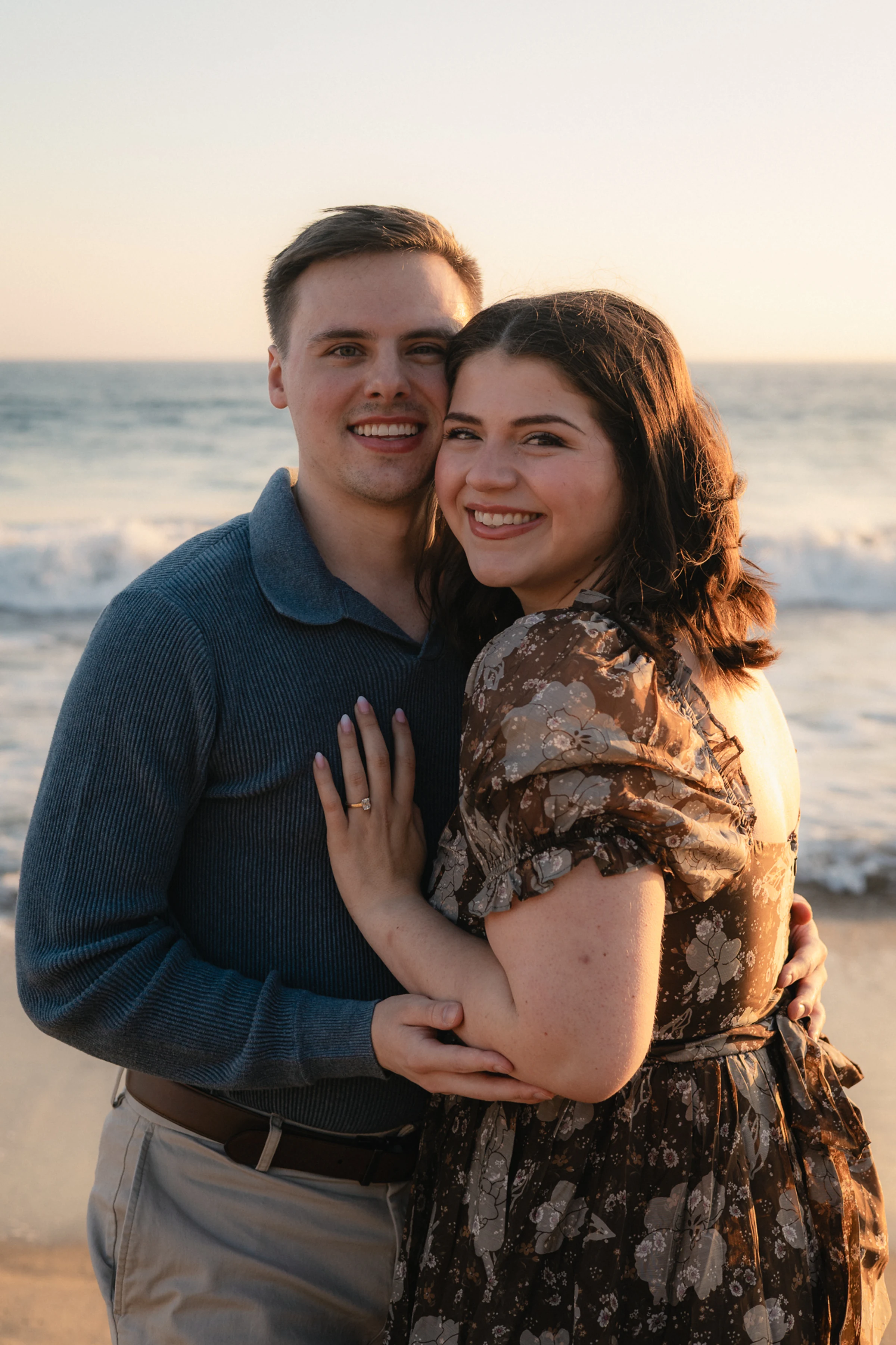 A smiling couple embraces on a beach at sunset. The man in a blue sweater and woman in a floral dress radiate warmth and joy against the ocean backdrop.