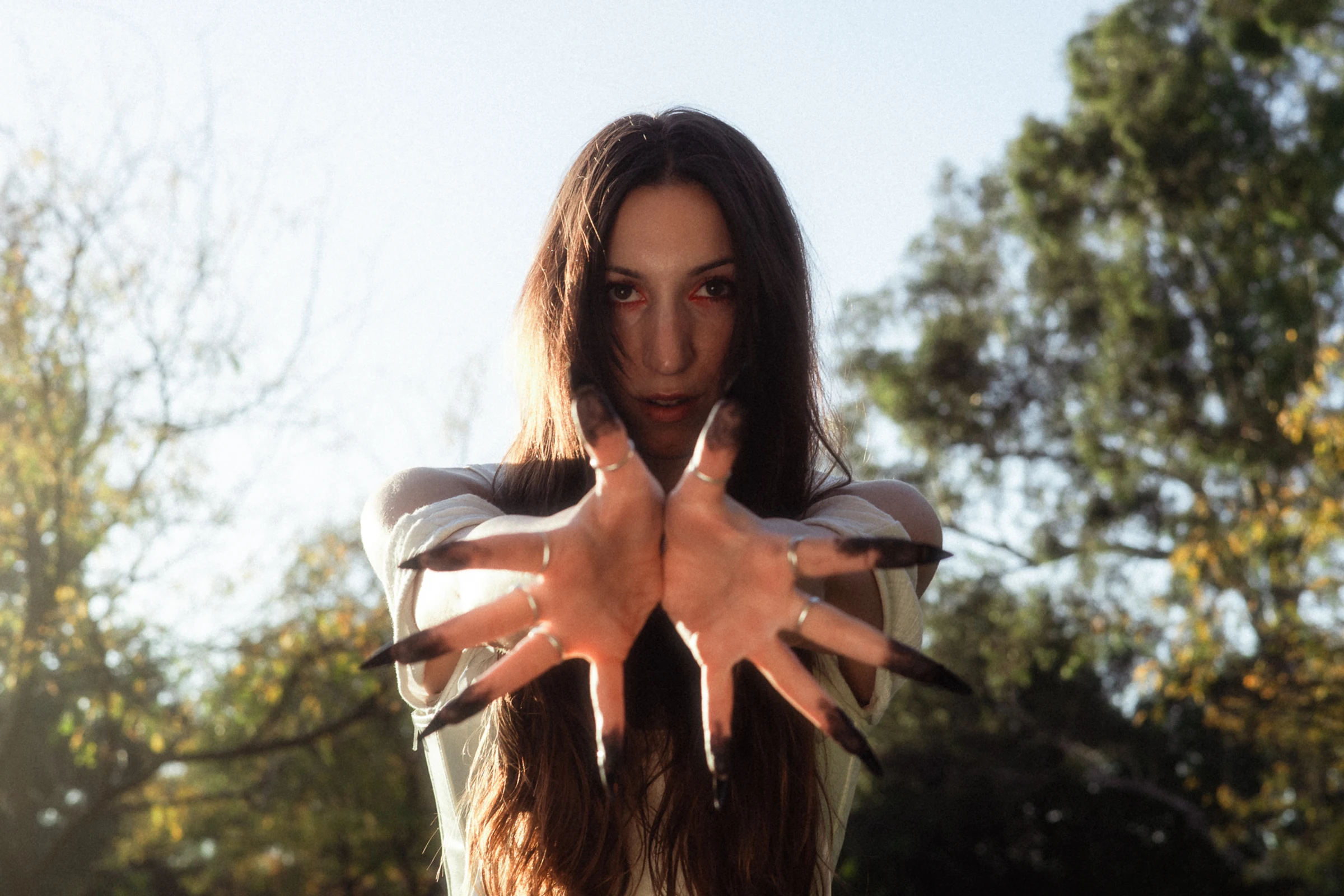 A woman with long brown hair extends her hands forward, featuring sharp, black fingernails. She stands outdoors under a clear sky, with trees in the background.